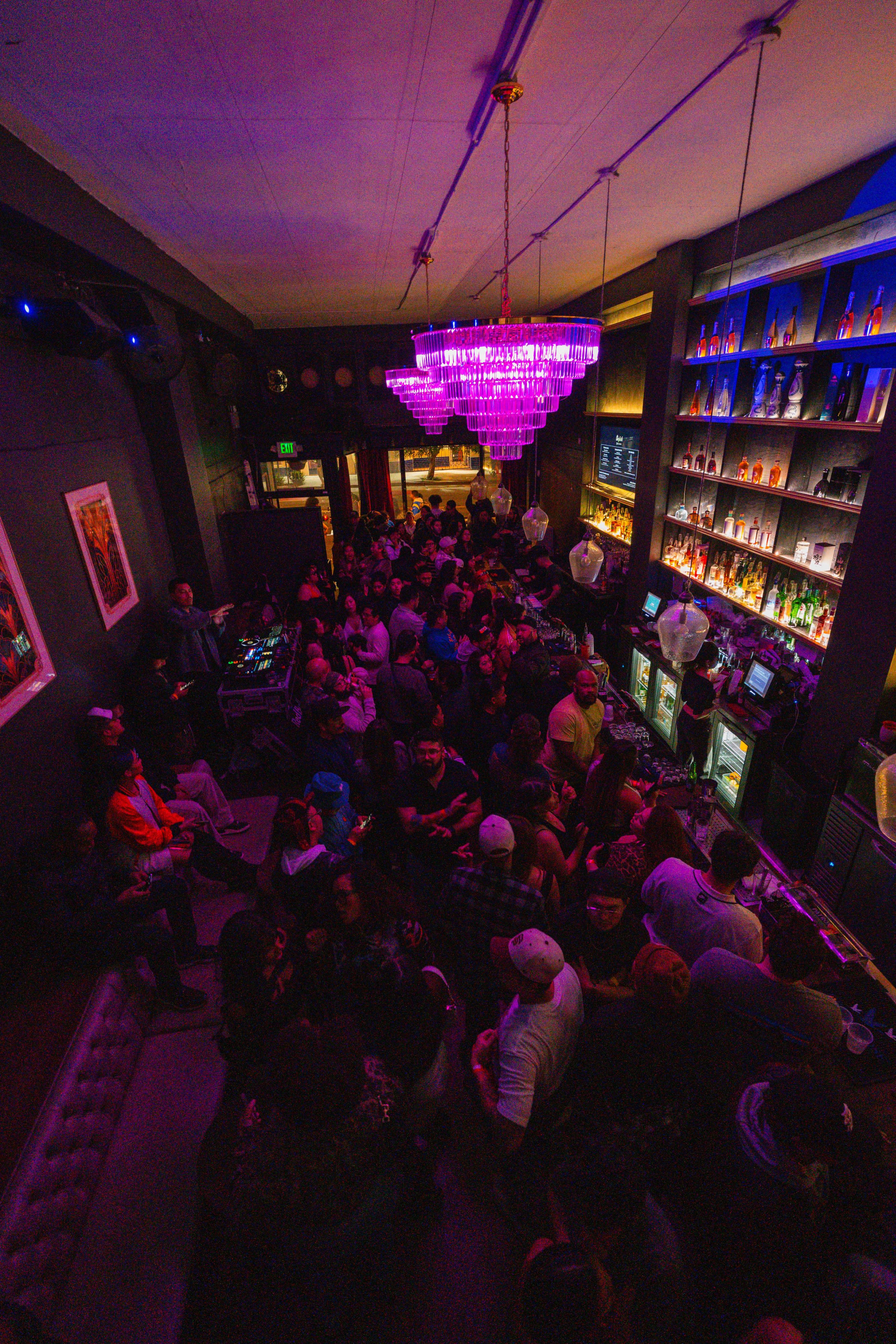 A large crowd mingles inside a dimly lit bar featuring a vibrant chandelier and shelves filled with colorful bottles.