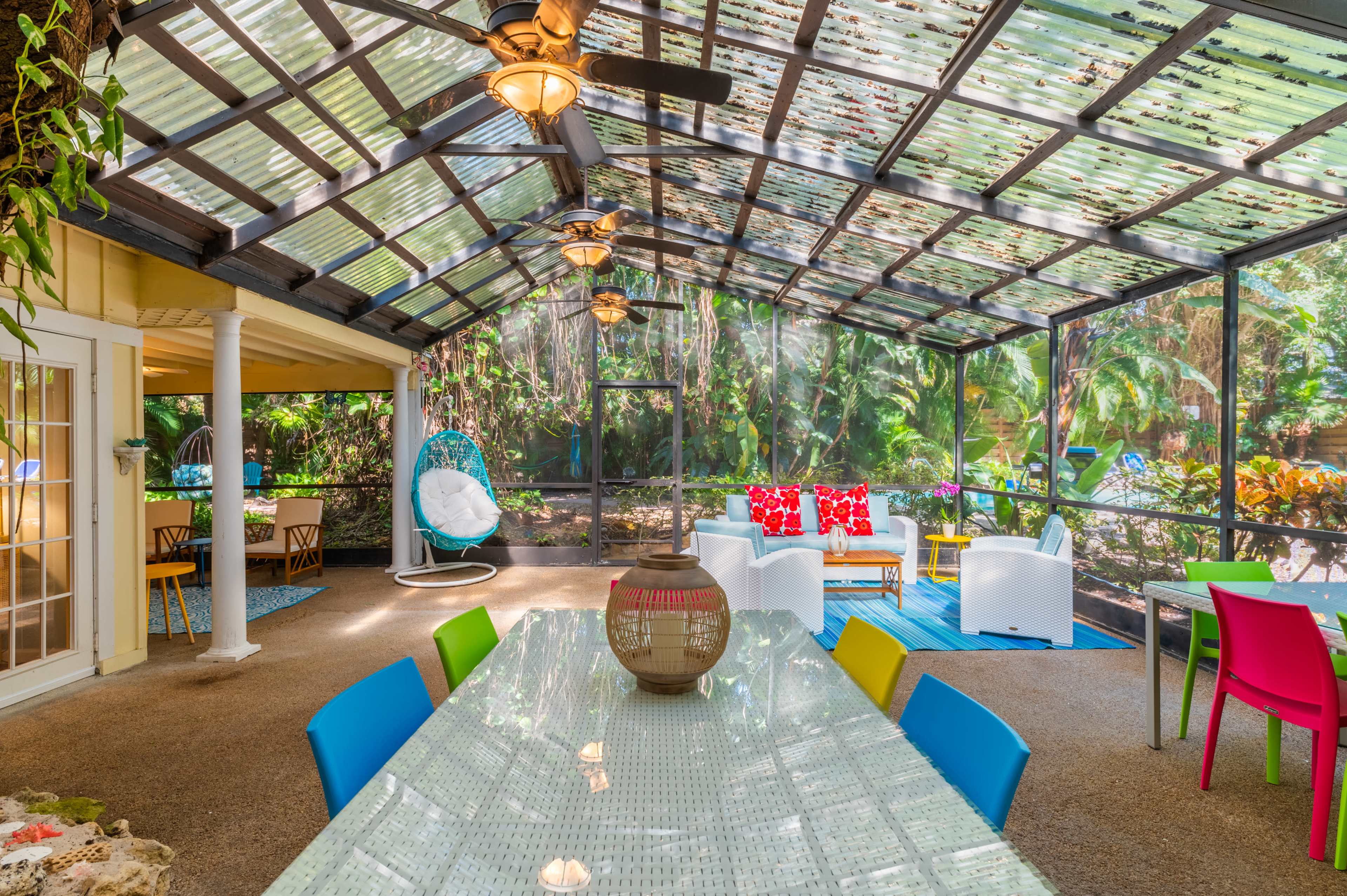 The image shows a spacious screened patio with colorful furniture, a large dining table, and lush greenery visible through the screens.