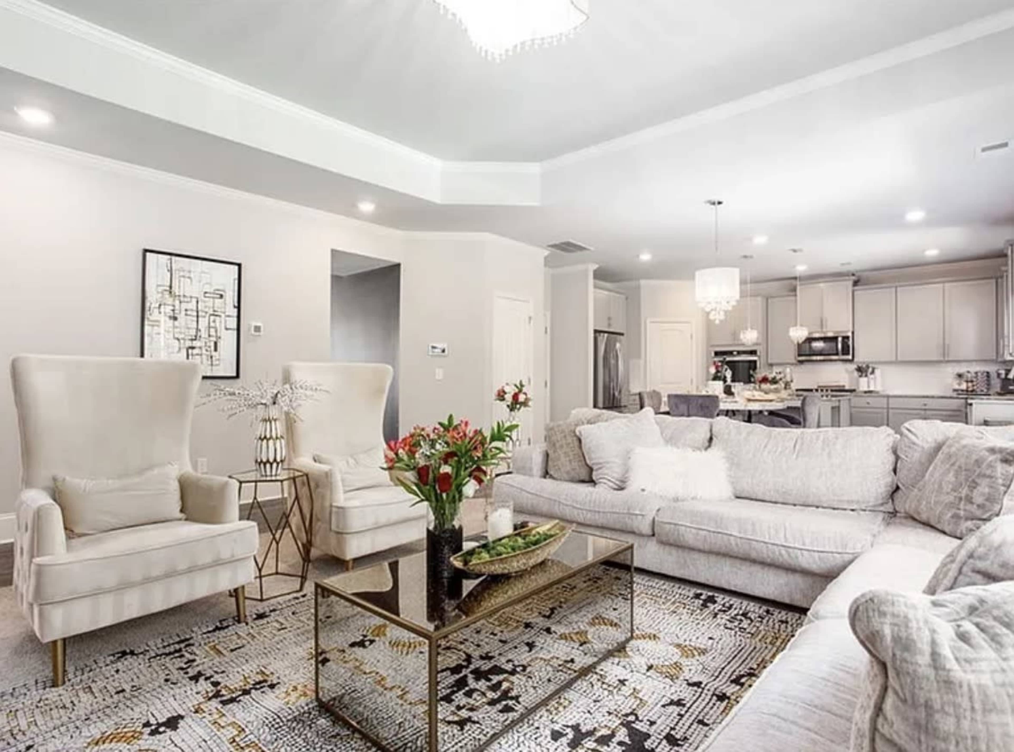 The image shows a modern living room featuring two wingback chairs, a plush sofa, a glass coffee table, and a patterned area rug.
