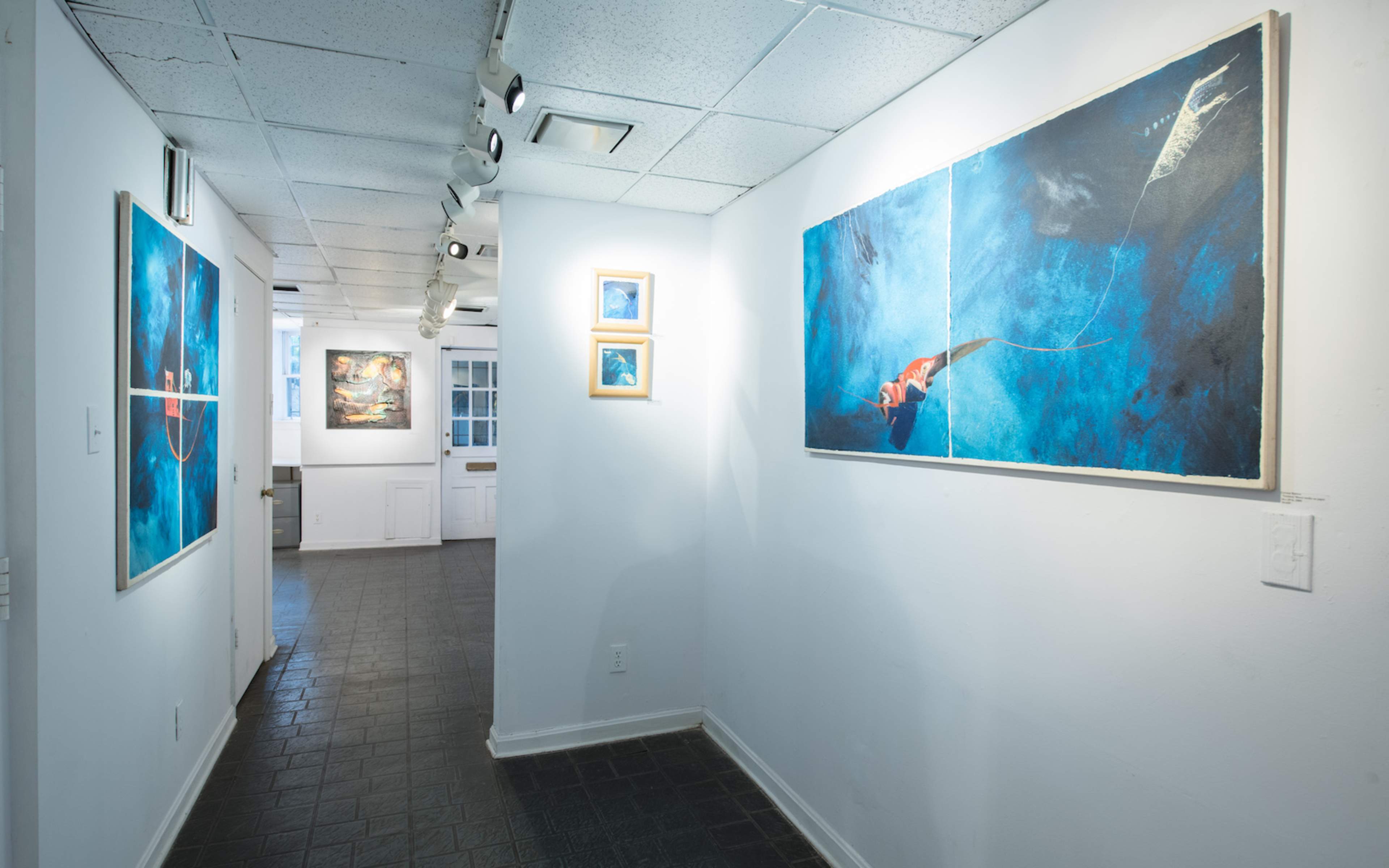 A narrow hallway within an art gallery, featuring two large paintings on the walls and smaller framed artworks displayed nearby.