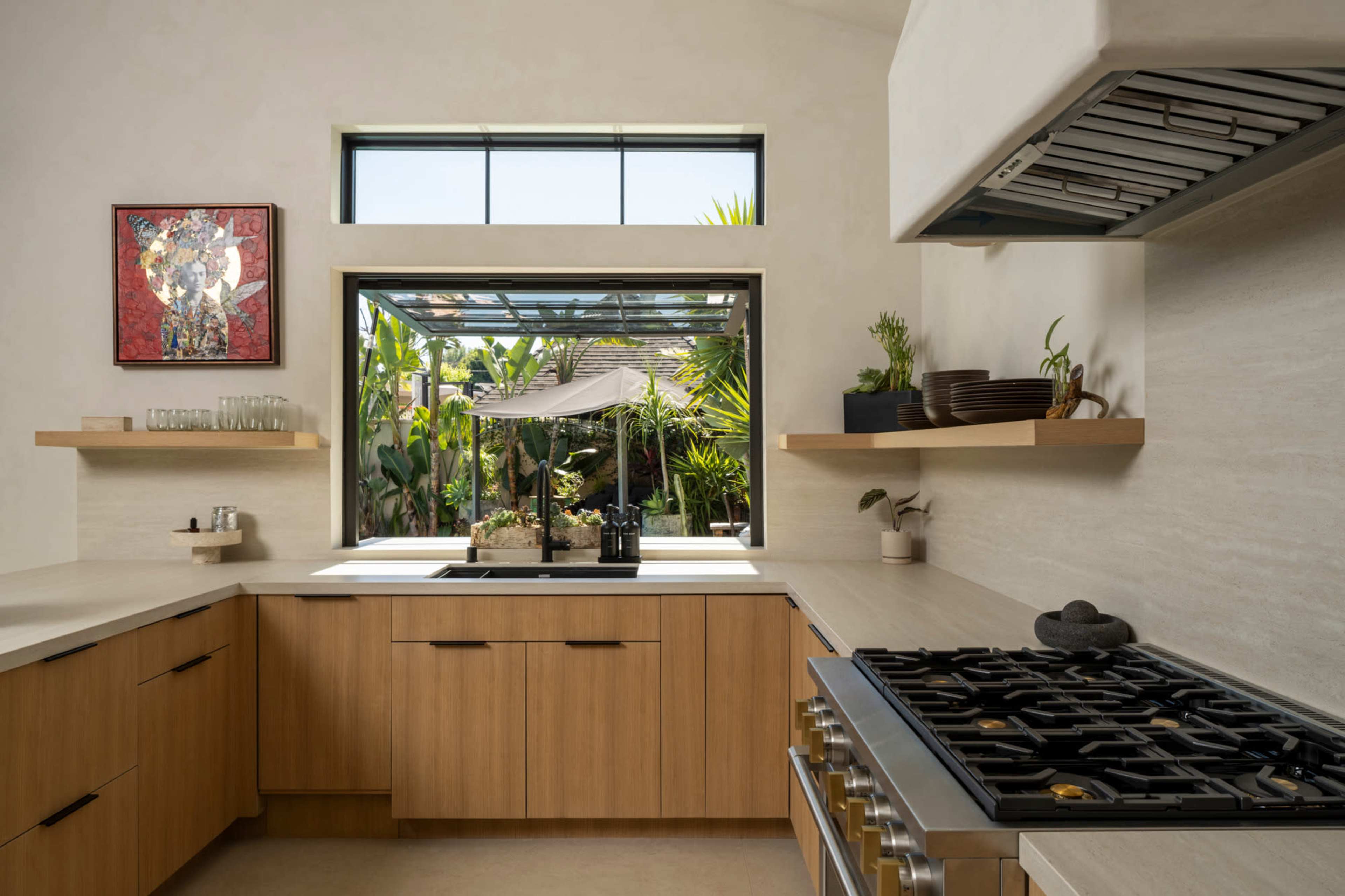 The image shows a modern kitchen with wooden cabinets, a large gas stove, and a window that opens to a view of greenery outside.
