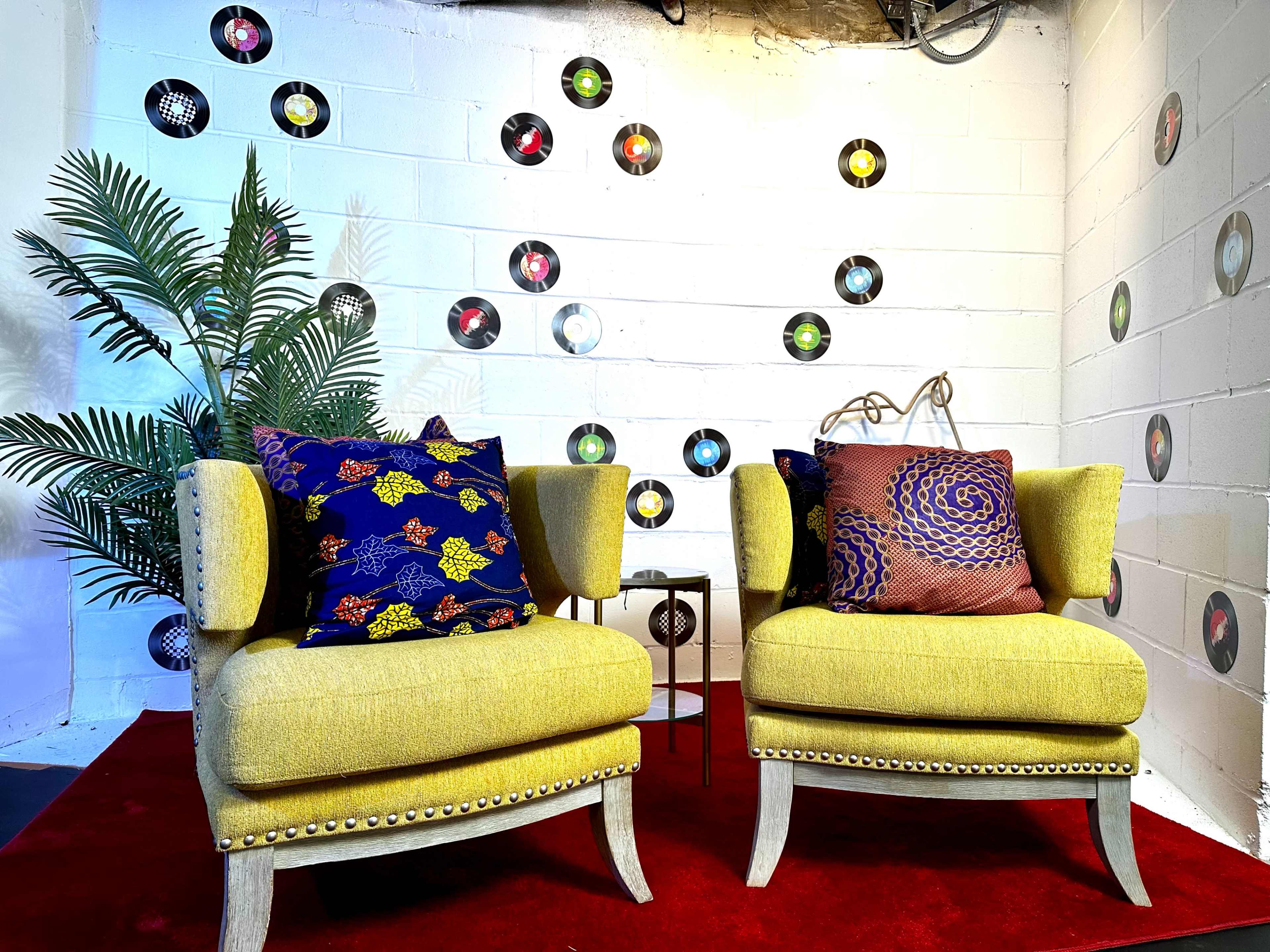 Two yellow chairs with decorative pillows are placed on a red carpet against a white wall decorated with vinyl records, alongside a small table and a potted plant.