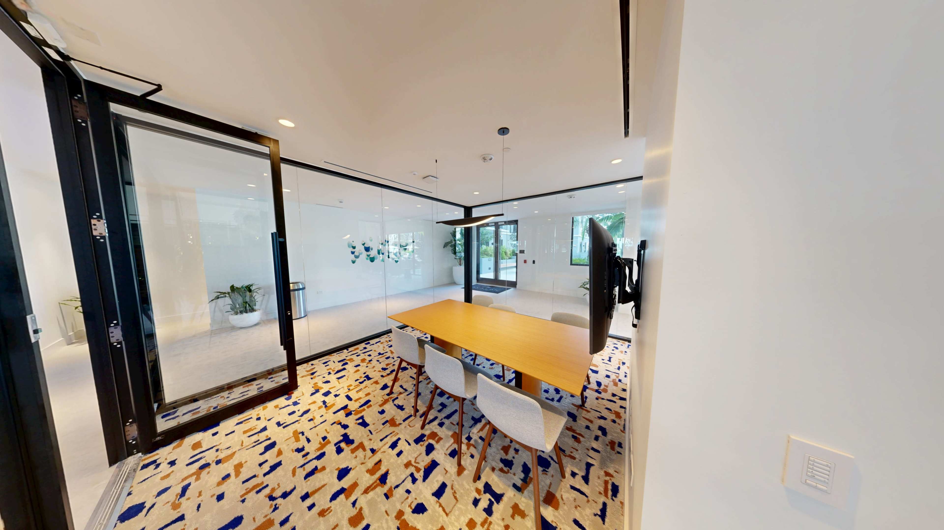 A modern conference room features a wooden table surrounded by chairs, with glass walls and a patterned carpet.