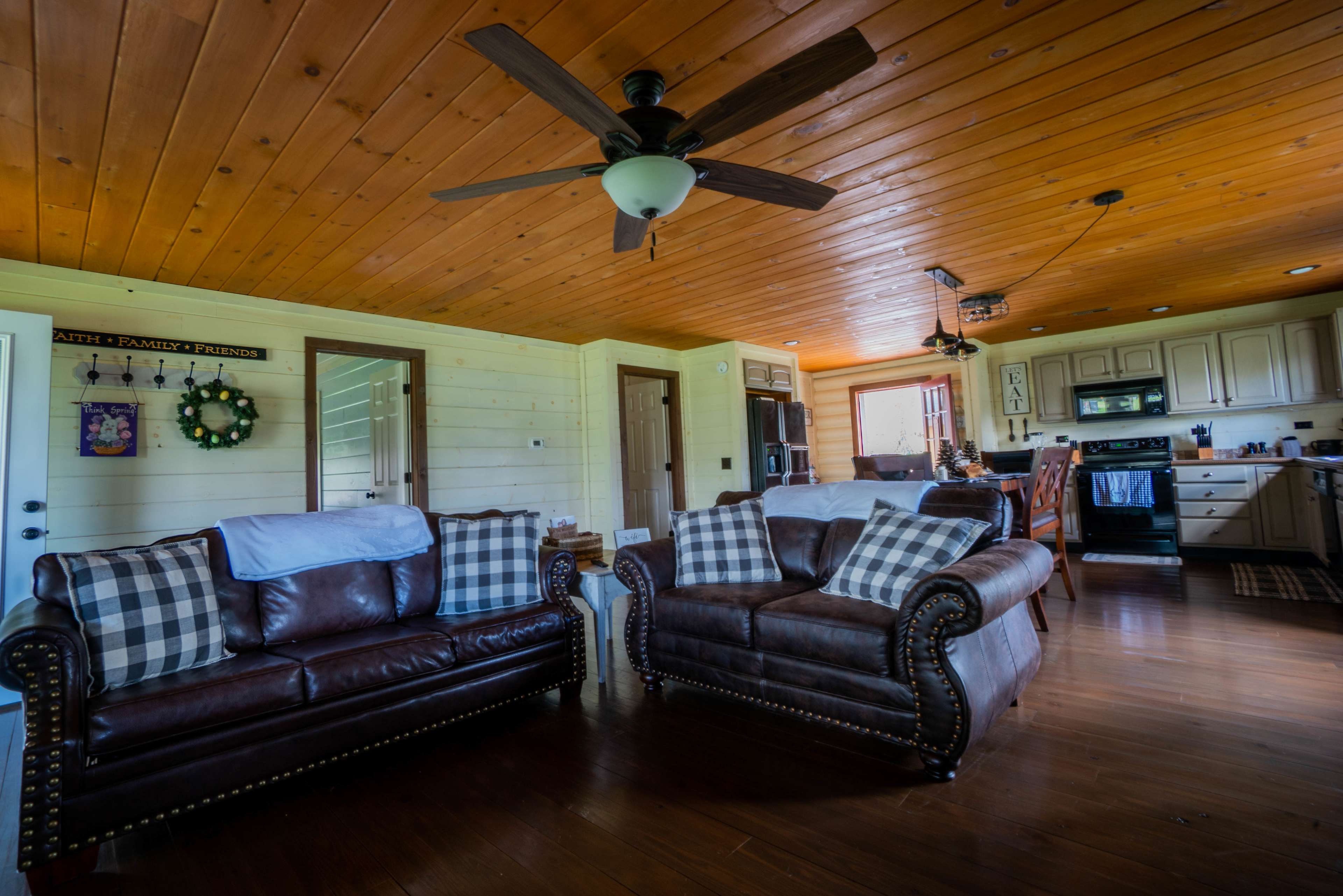 The image shows a cozy living room with two brown leather sofas adorned with checkered pillows, wooden ceiling, and an open kitchen area in the background.