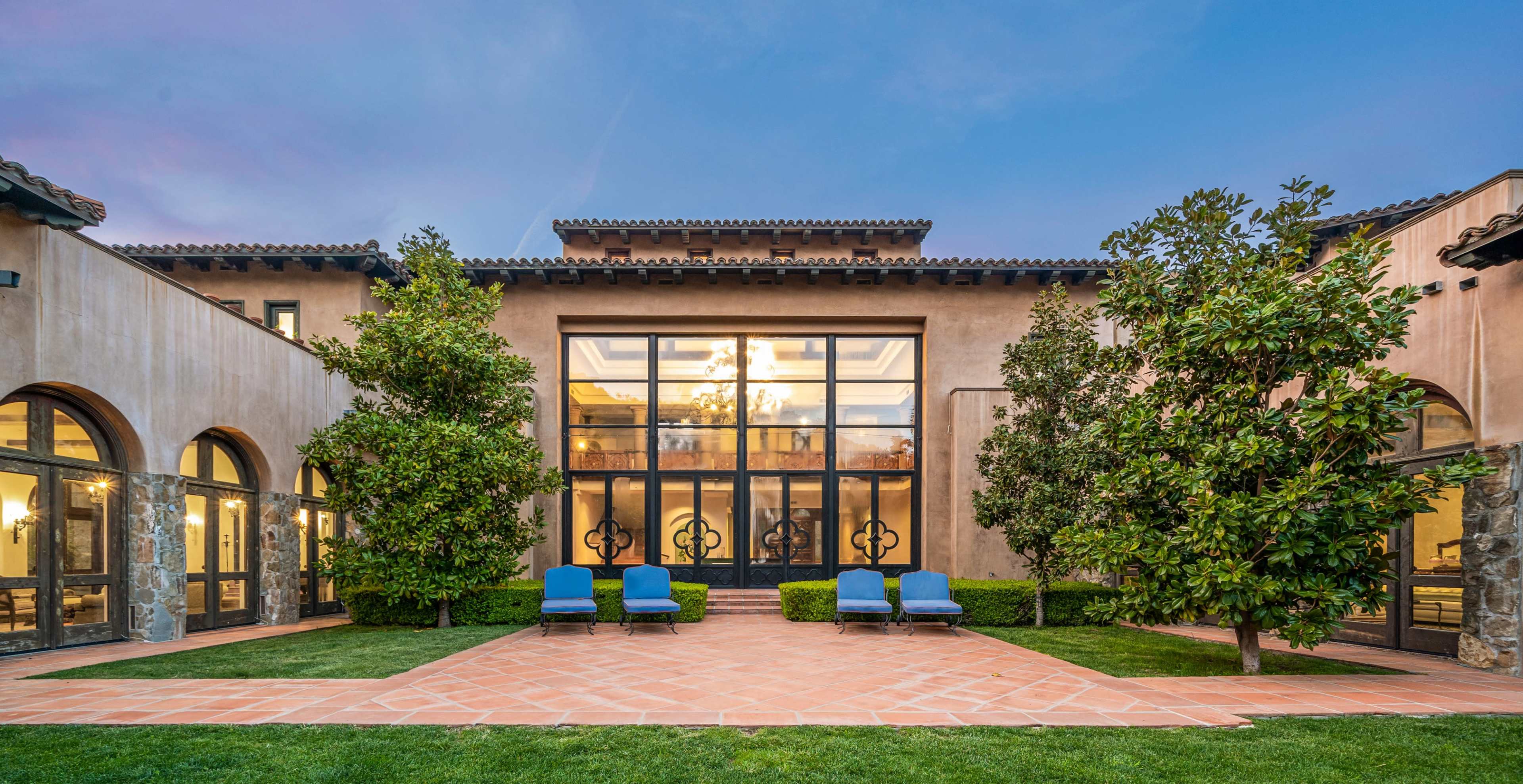 The image shows an elegant exterior view of a large house featuring a prominent glass-walled section, flanked by manicured greenery and outdoor lounge chairs arranged on a tiled path.