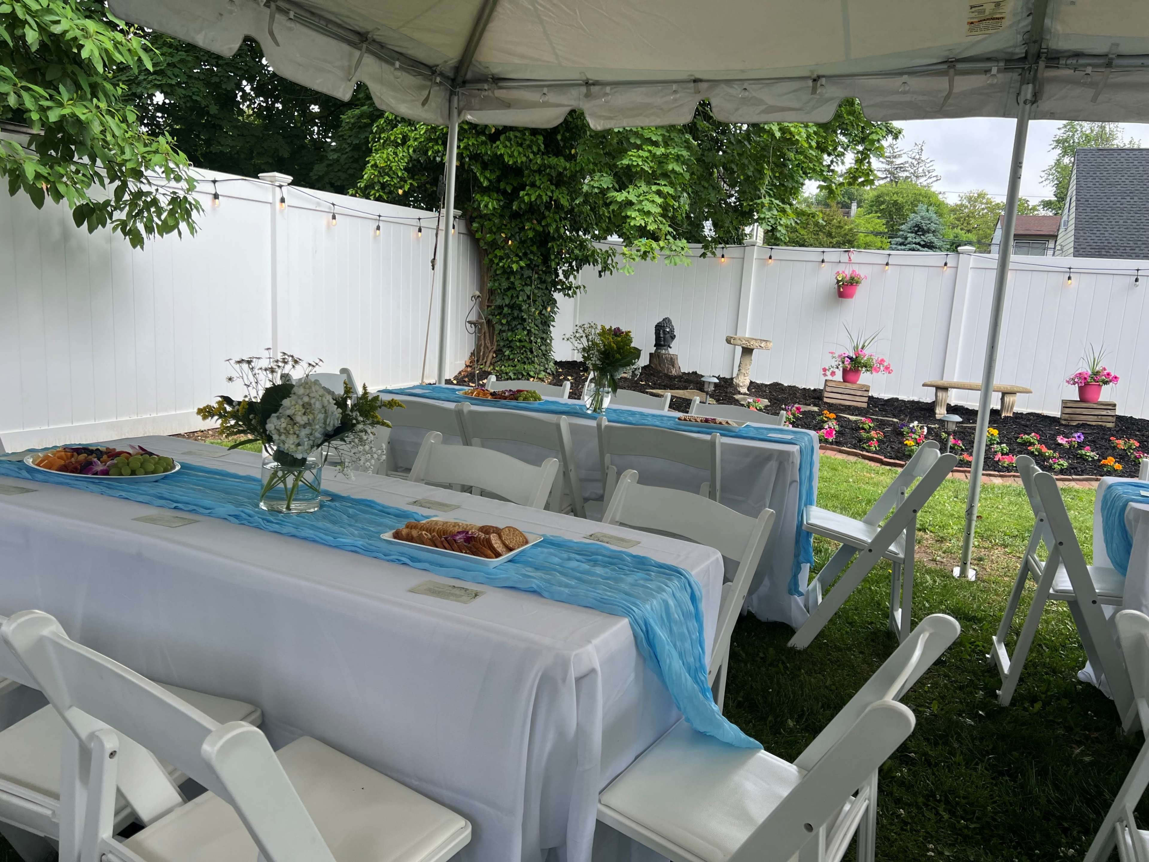 A tented outdoor space features neatly arranged tables with white tablecloths and blue runners, surrounded by a garden with flowers and decorative elements.