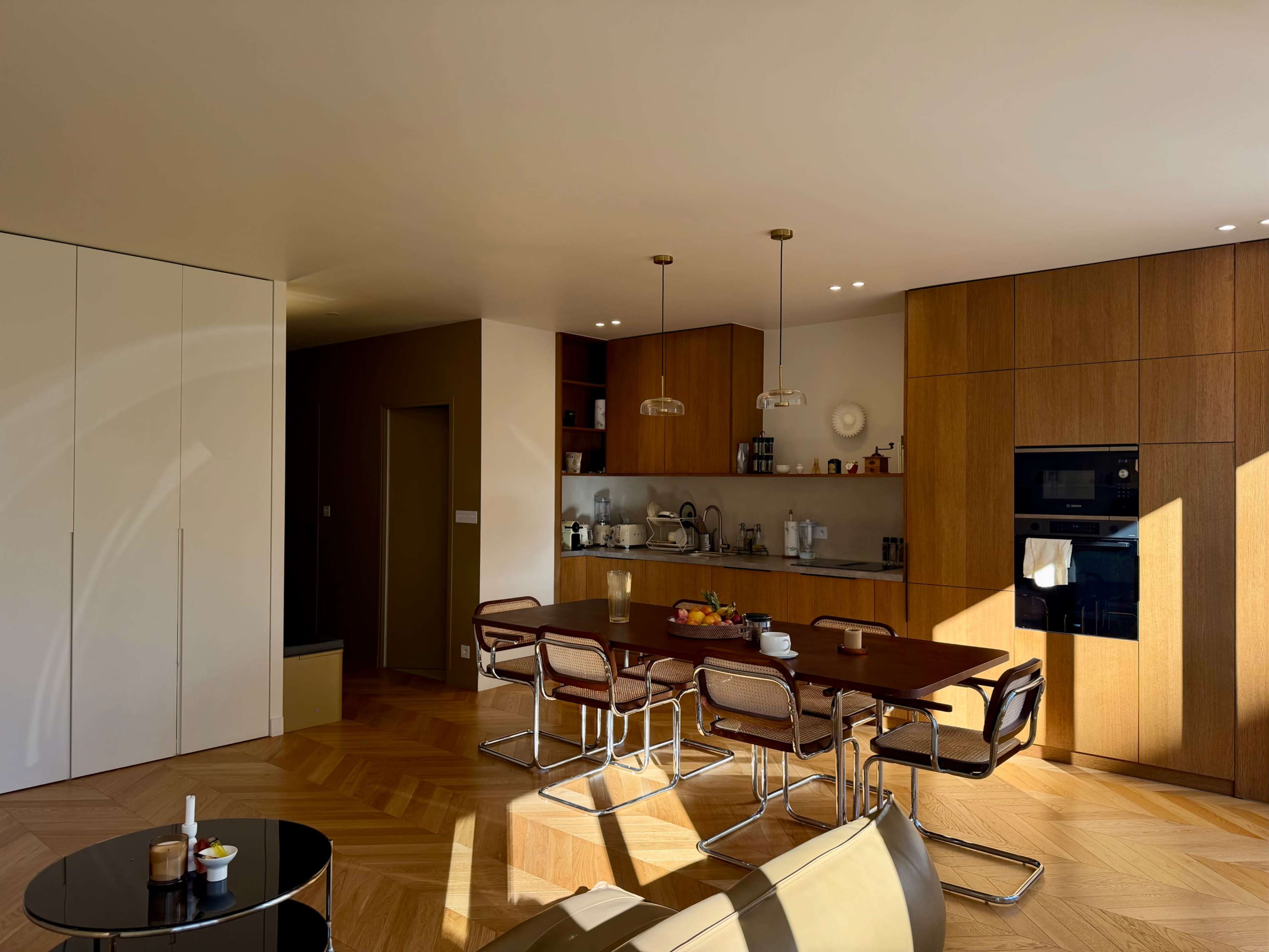 The image shows a modern kitchen and dining area with wooden cabinetry, a large table with chairs, and large windows allowing natural light to fill the space.