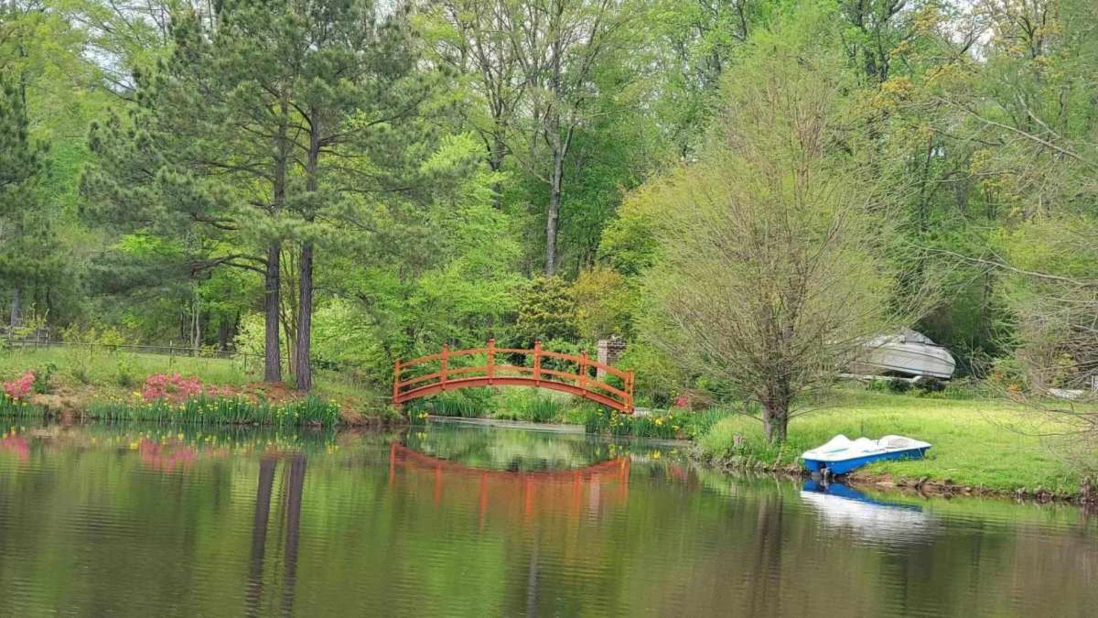A red wooden bridge crosses a calm pond surrounded by greenery and flowers, with a small boat tied to a dock nearby.