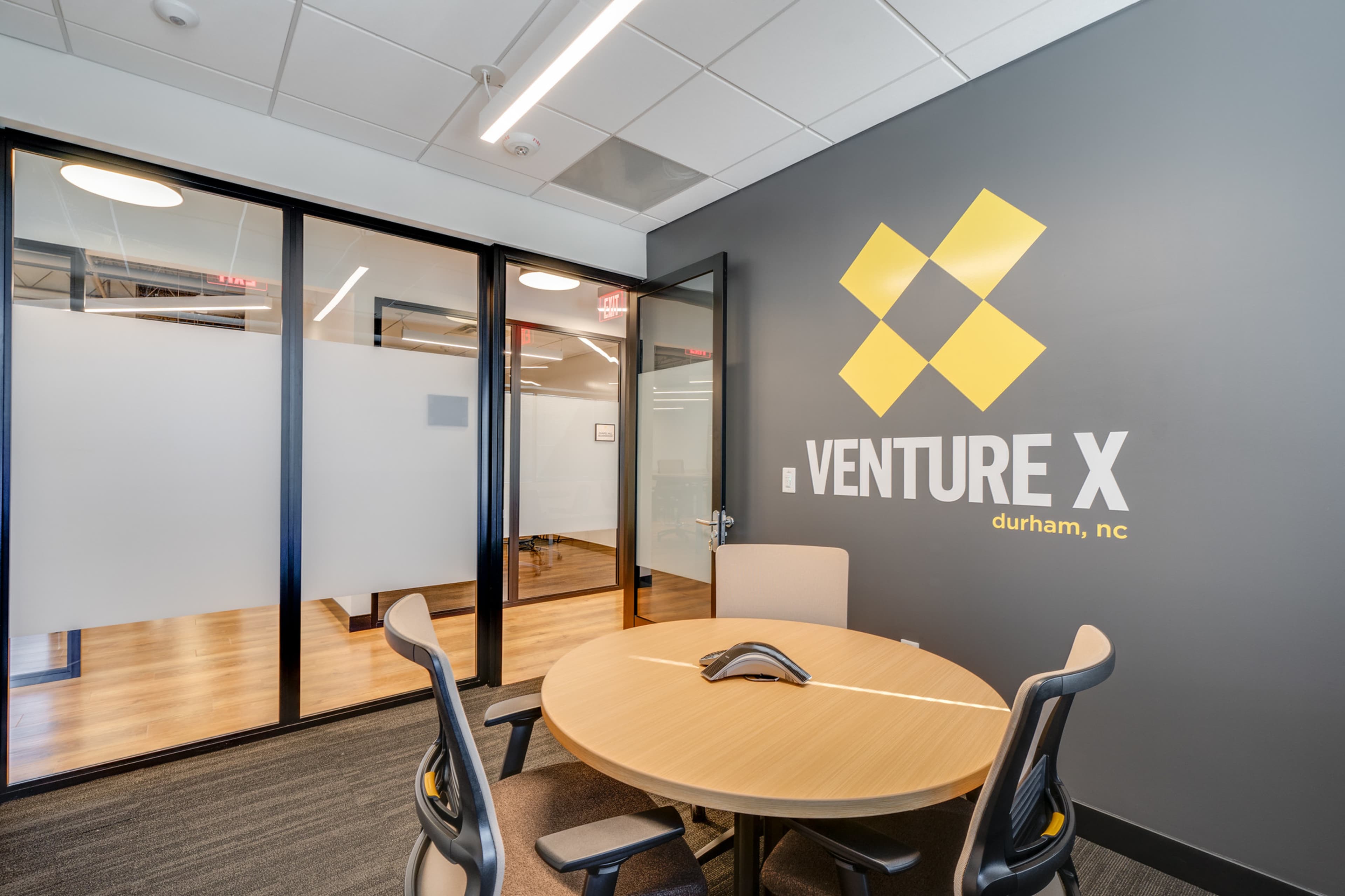 A modern office space with a round table and chairs, featuring a wall with the logo "VENTURE X" and the location "durham, nc."