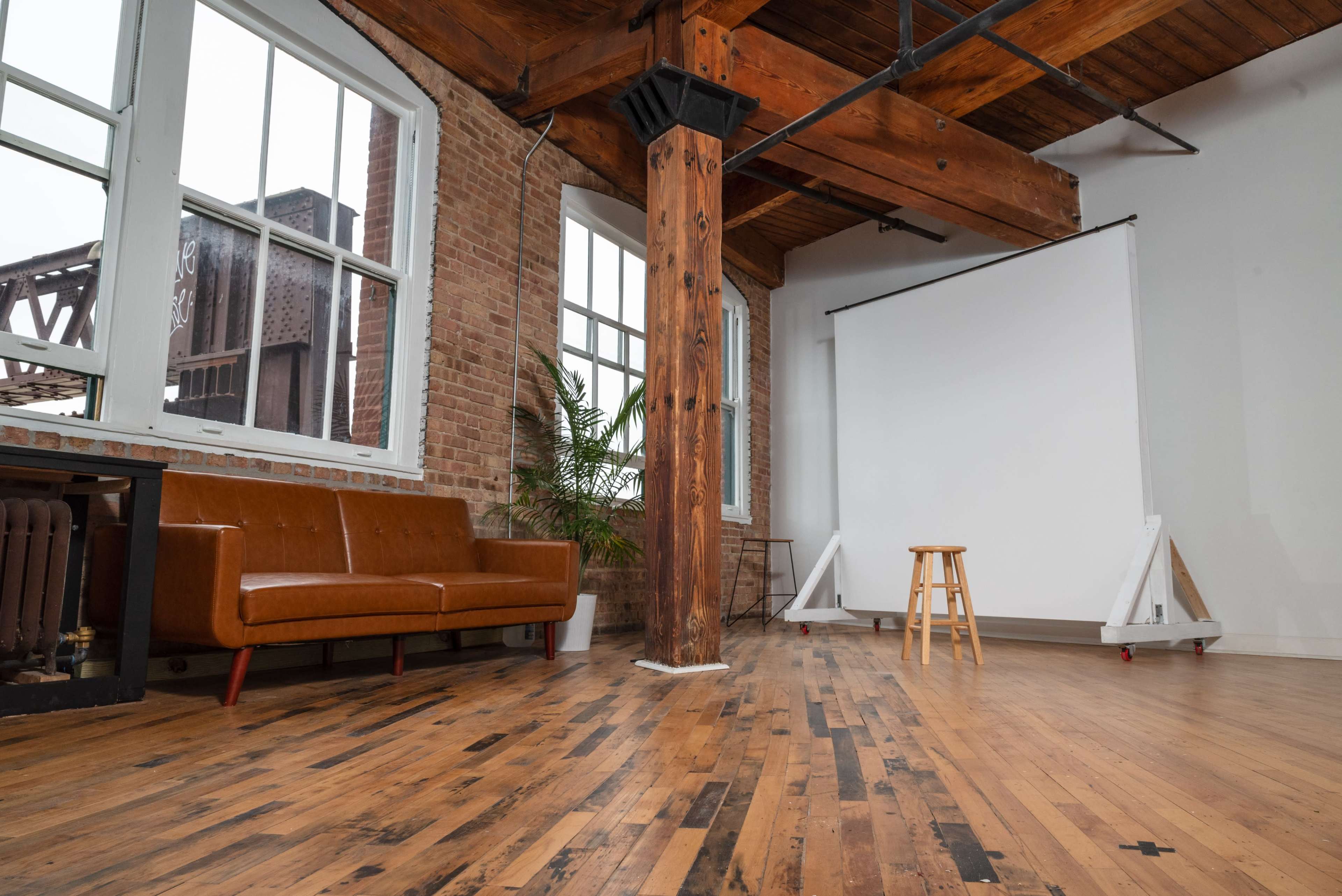 An industrial-style room with exposed brick walls, wooden beams, a brown sofa, and a white backdrop on a stool for photography.