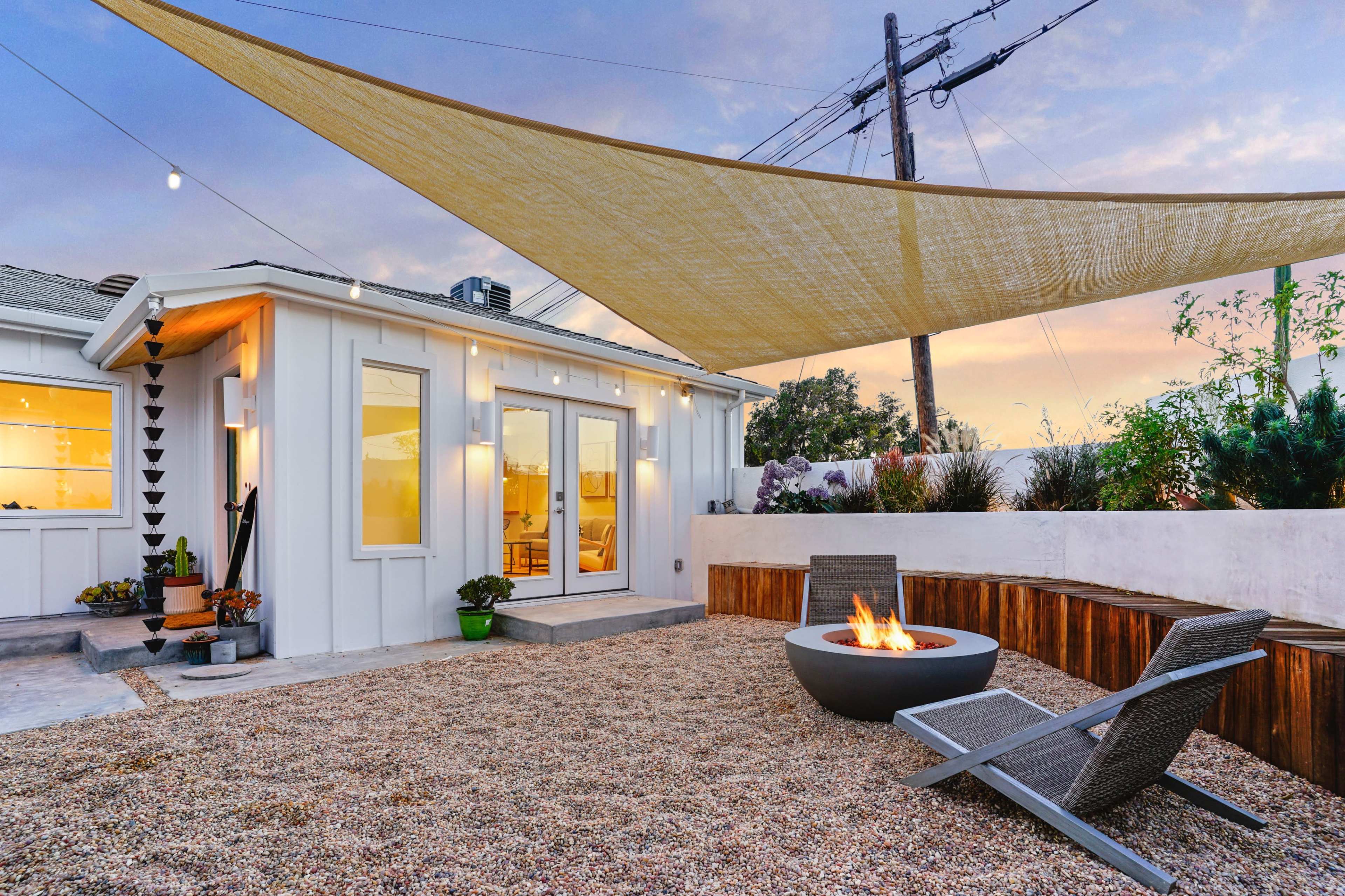 A modern outdoor space features a fire pit surrounded by gravel, lounge chairs, and a shaded area with a fabric canopy.