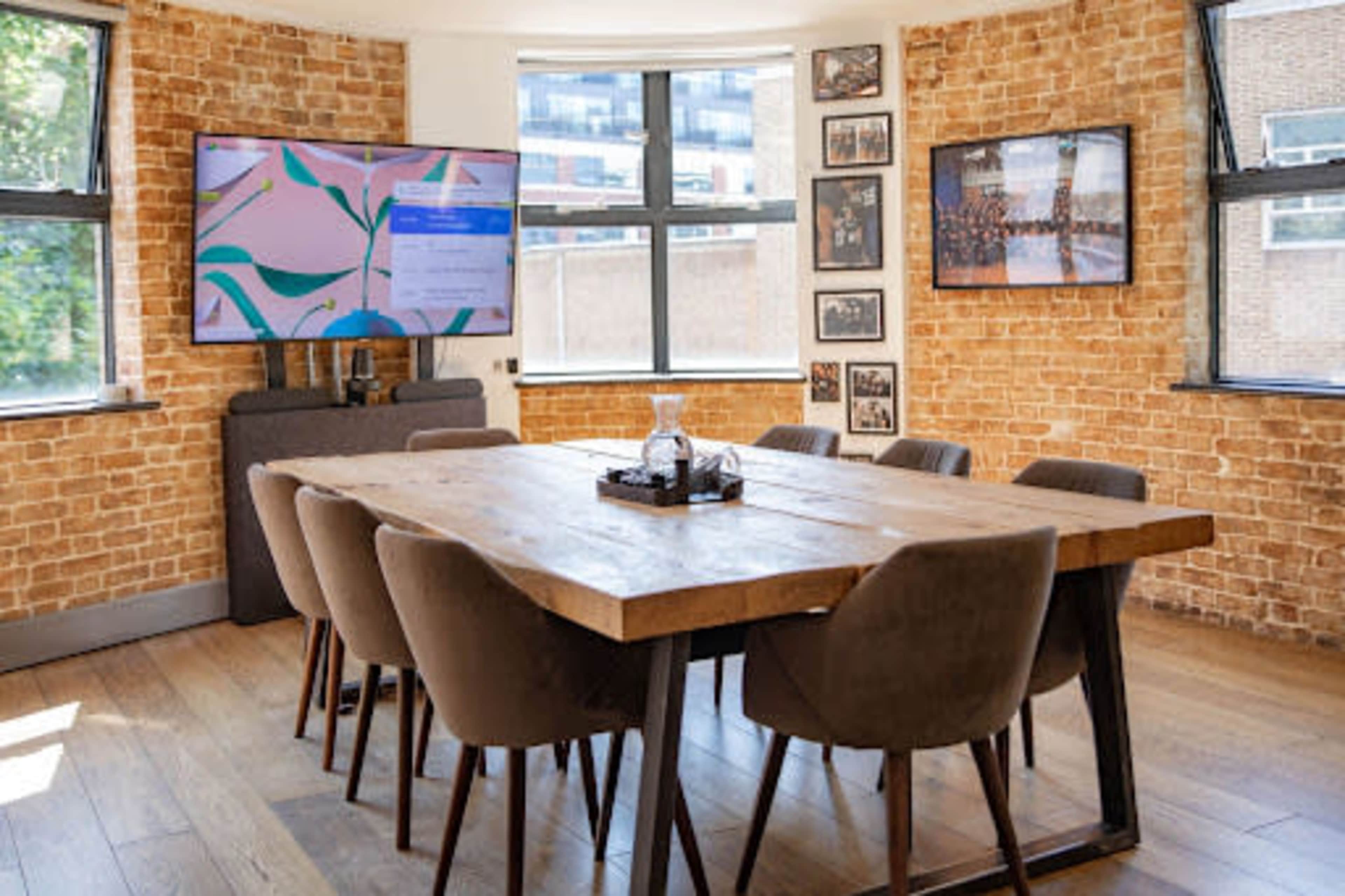 A modern conference room features a large wooden table surrounded by upholstered chairs, with a television mounted on the wall and brick accents.