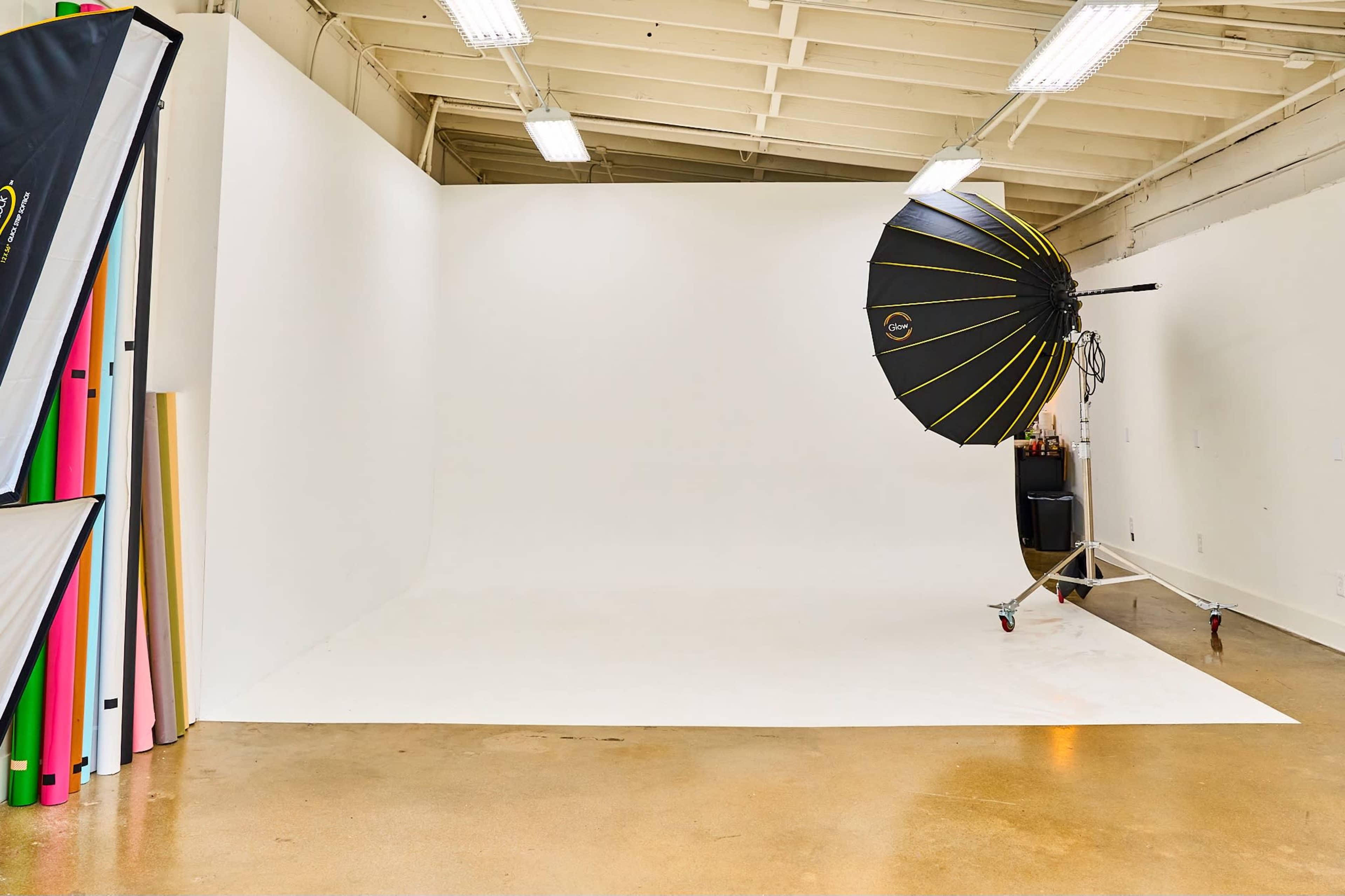 A photography studio with a white backdrop, an umbrella light, and a storage area for colorful backdrop rolls.