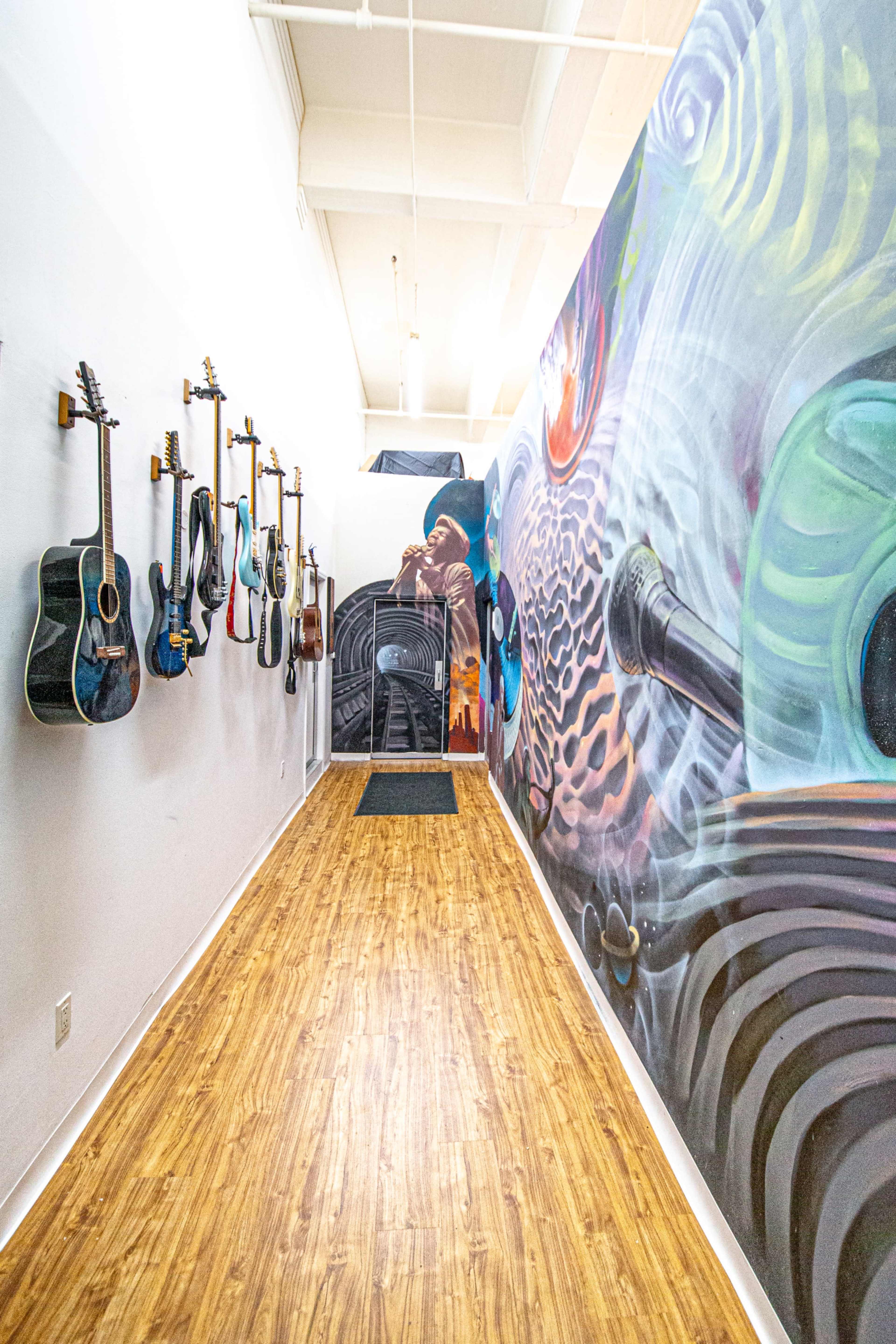The image features a hallway lined with various acoustic guitars hanging on the left side, accompanied by a colorful mural on the right wall depicting abstract designs.