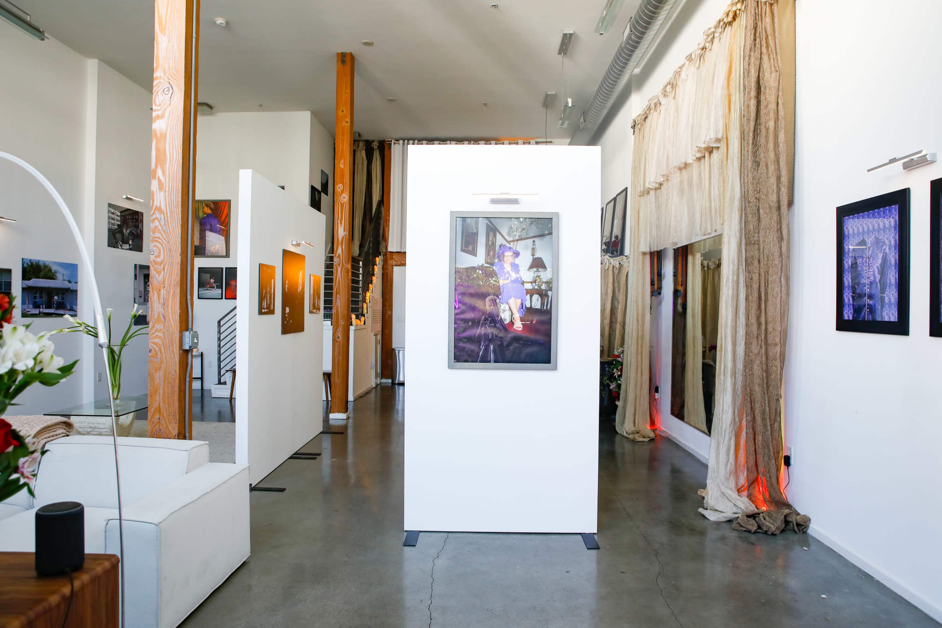 An art gallery interior with white walls displaying framed artwork, wooden beams, and a staircase in the background.