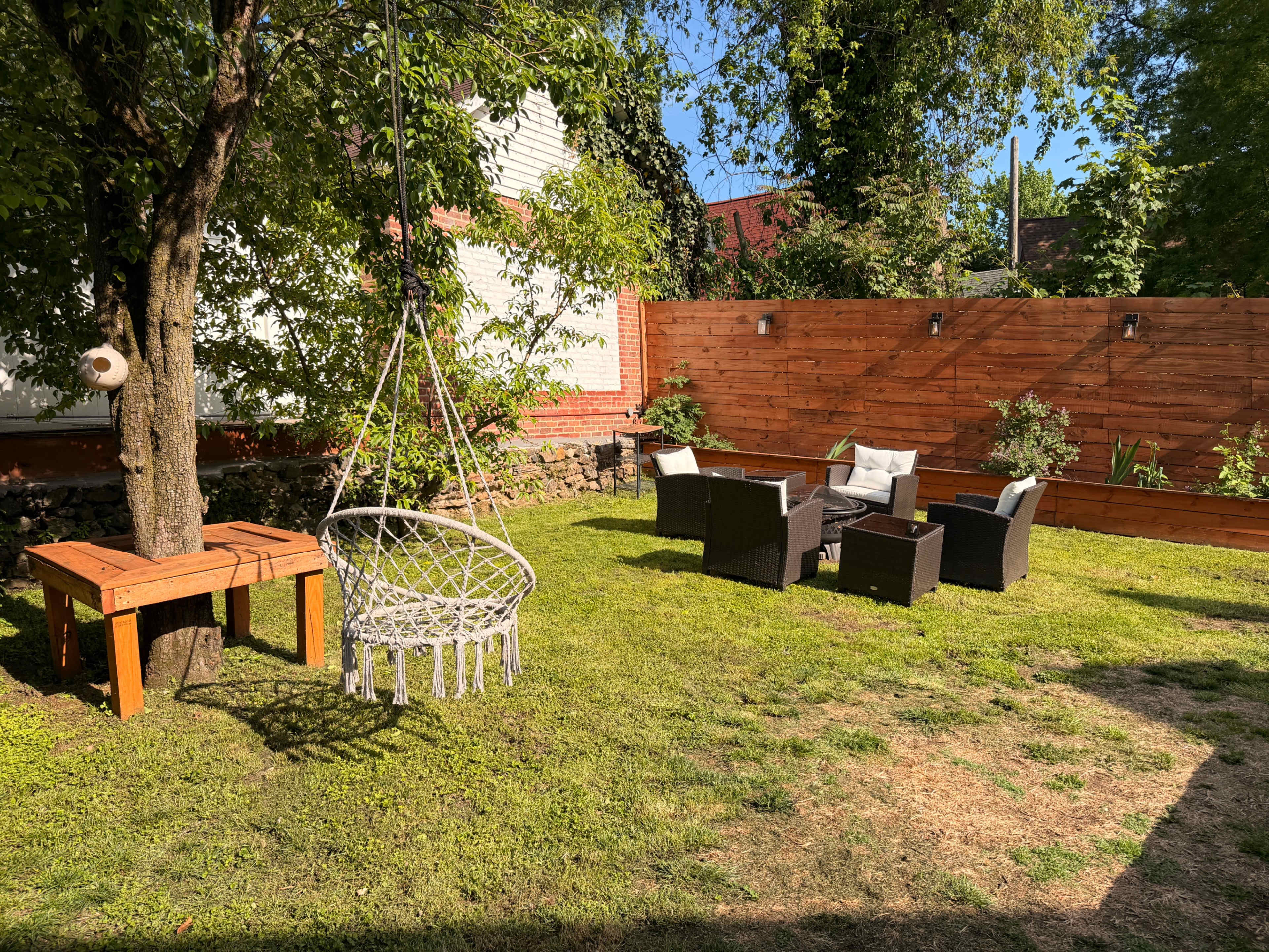 The image shows a backyard with a swing hanging from a tree, a wooden table, and a set of wicker chairs arranged on a green lawn surrounded by wooden fencing.