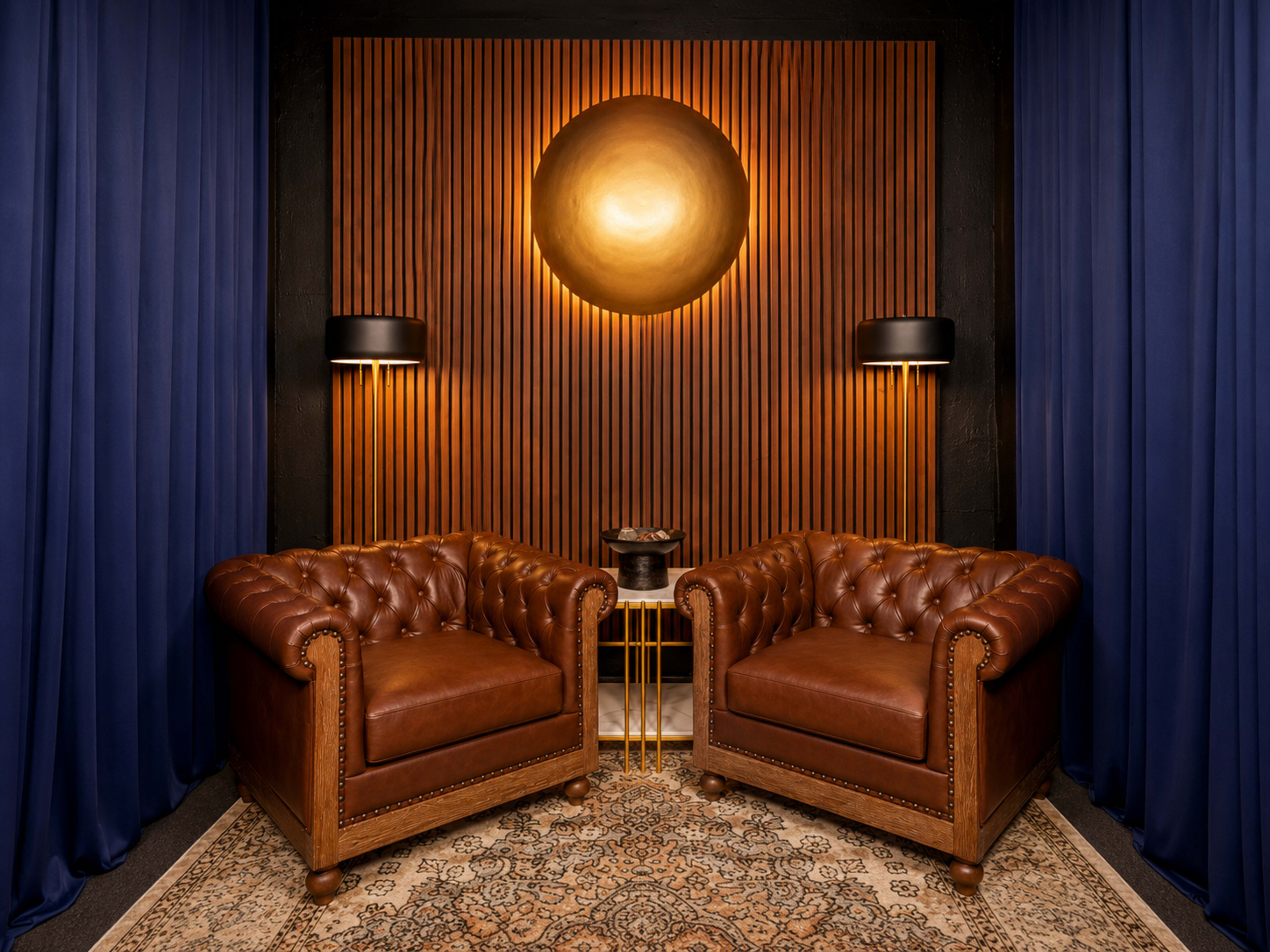 The image features two brown leather tufted chairs positioned against a wooden slatted wall and illuminated by circular golden wall art and two modern black lamps.