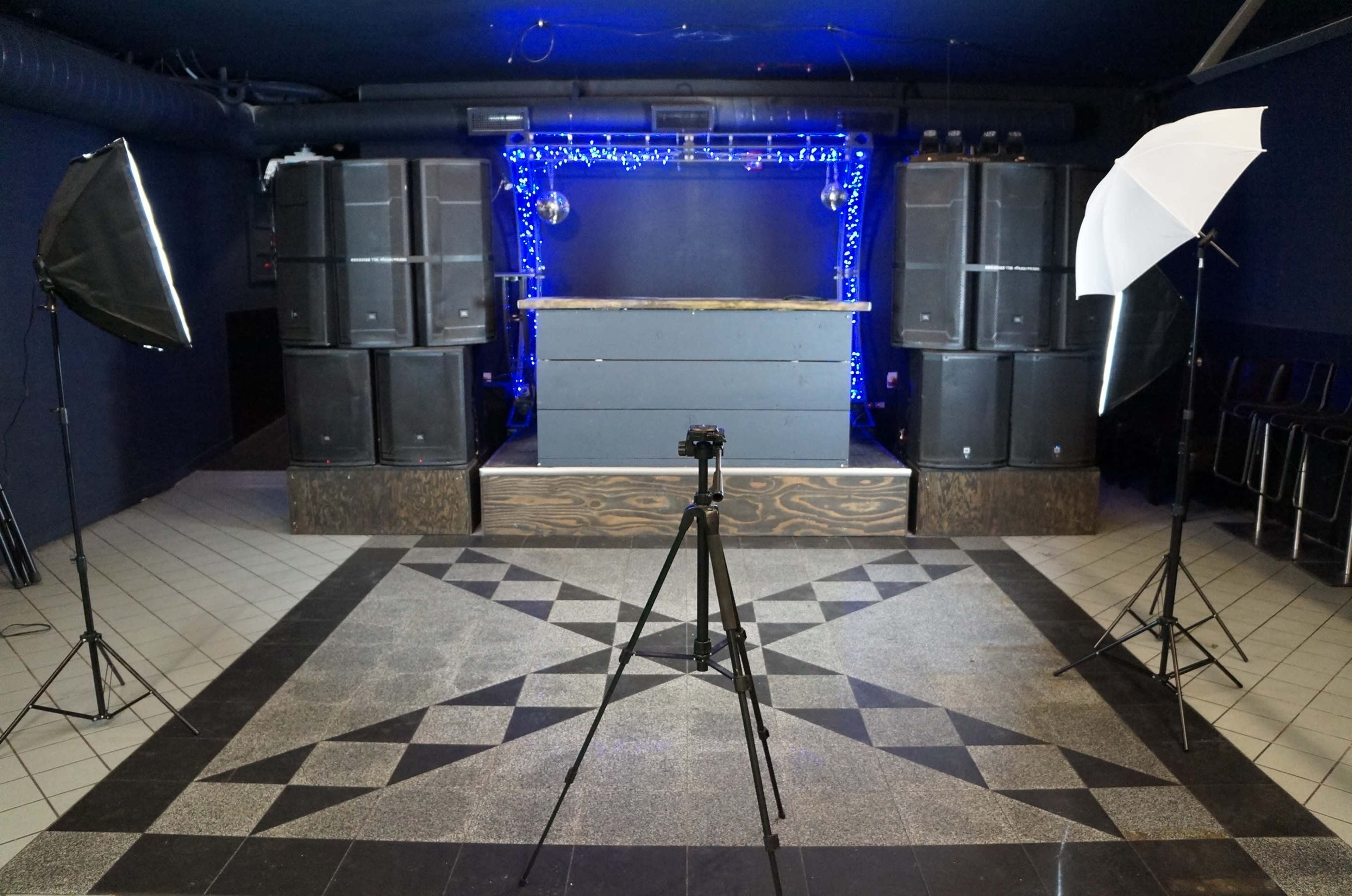 The scene features a photography setup in a dimly lit venue with a DJ booth, speakers, and lighting equipment arranged in front of a patterned floor.