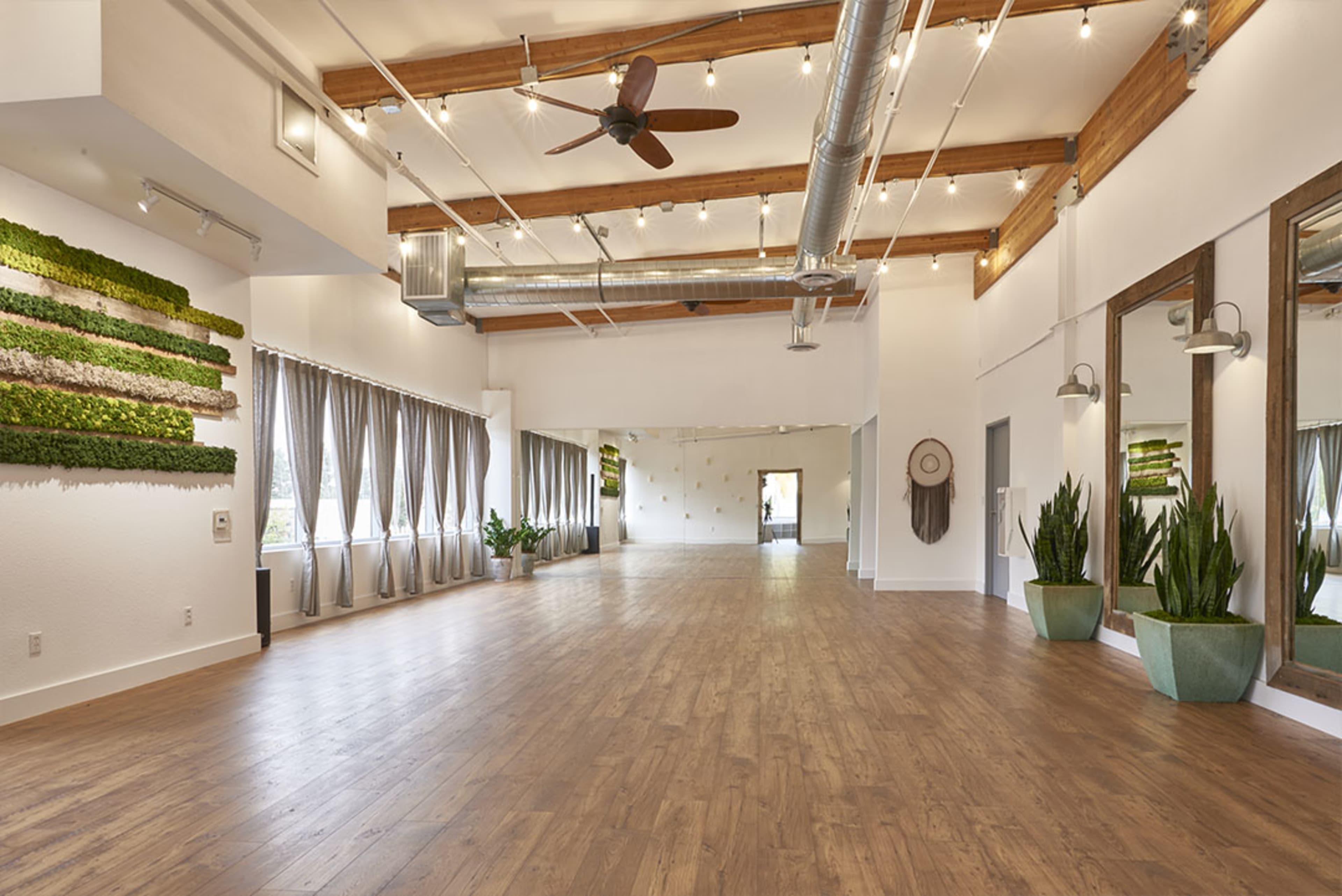 The image shows a spacious, well-lit interior with wooden flooring, large mirrors, and greenery accents along the walls.