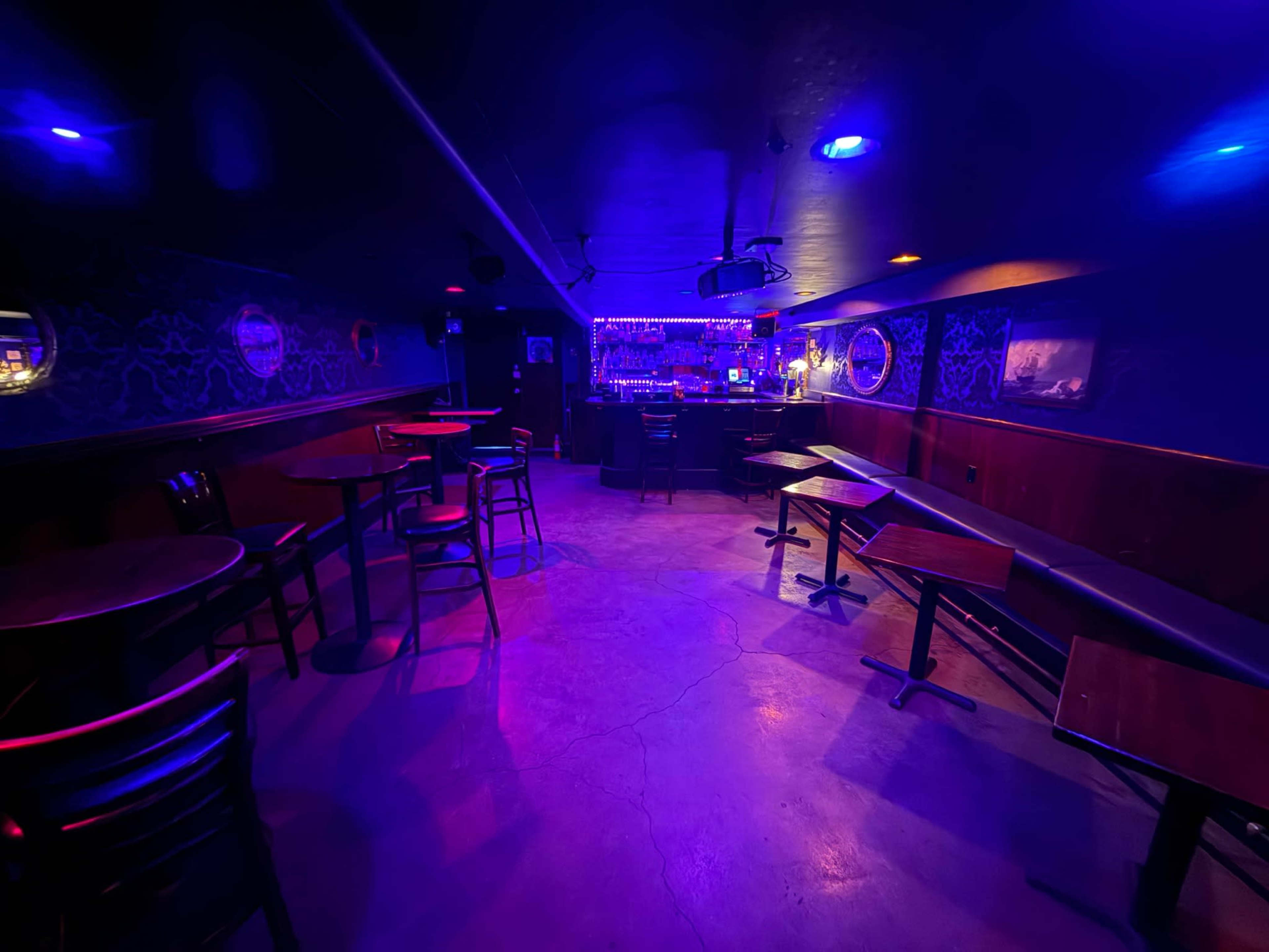 The image shows a dimly lit bar interior with empty tables and a brightly illuminated bar counter at the back, surrounded by walls adorned with decorative motifs.
