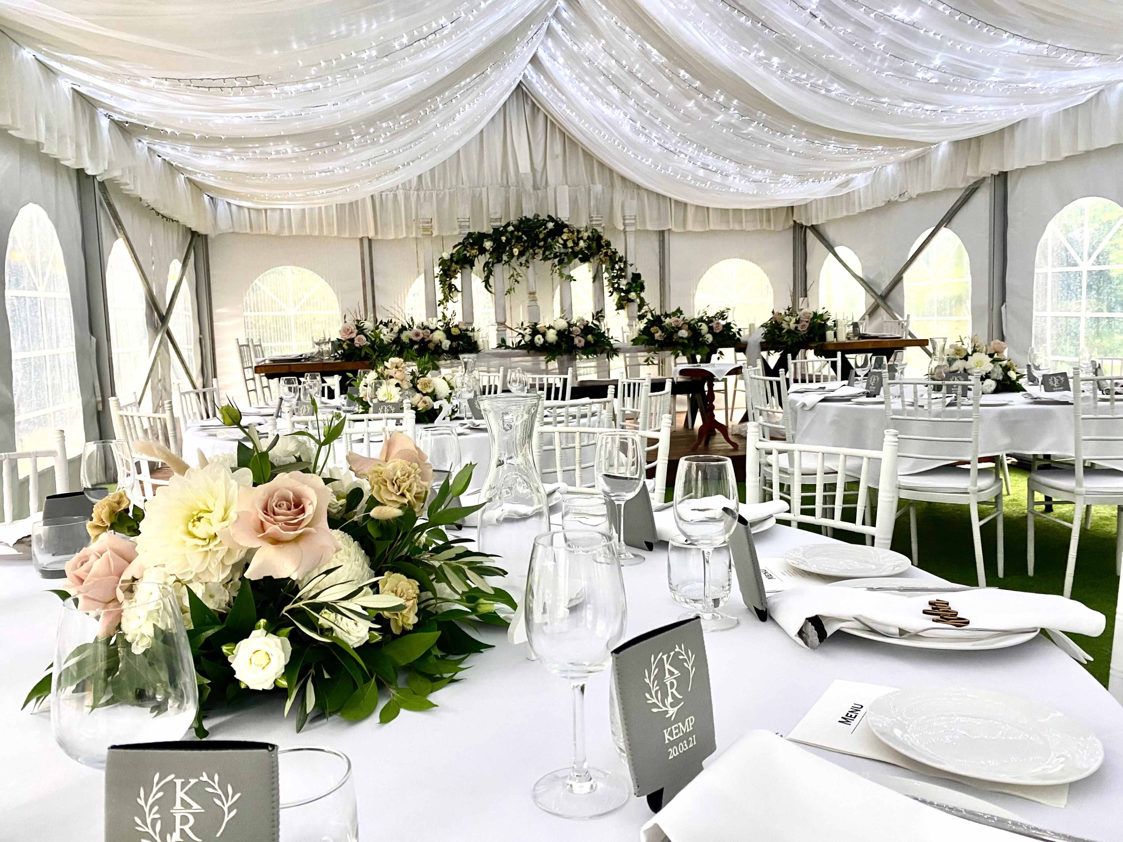 A decorated tent with tables set for a formal event, featuring floral centerpieces, elegant tableware, and a floral backdrop.