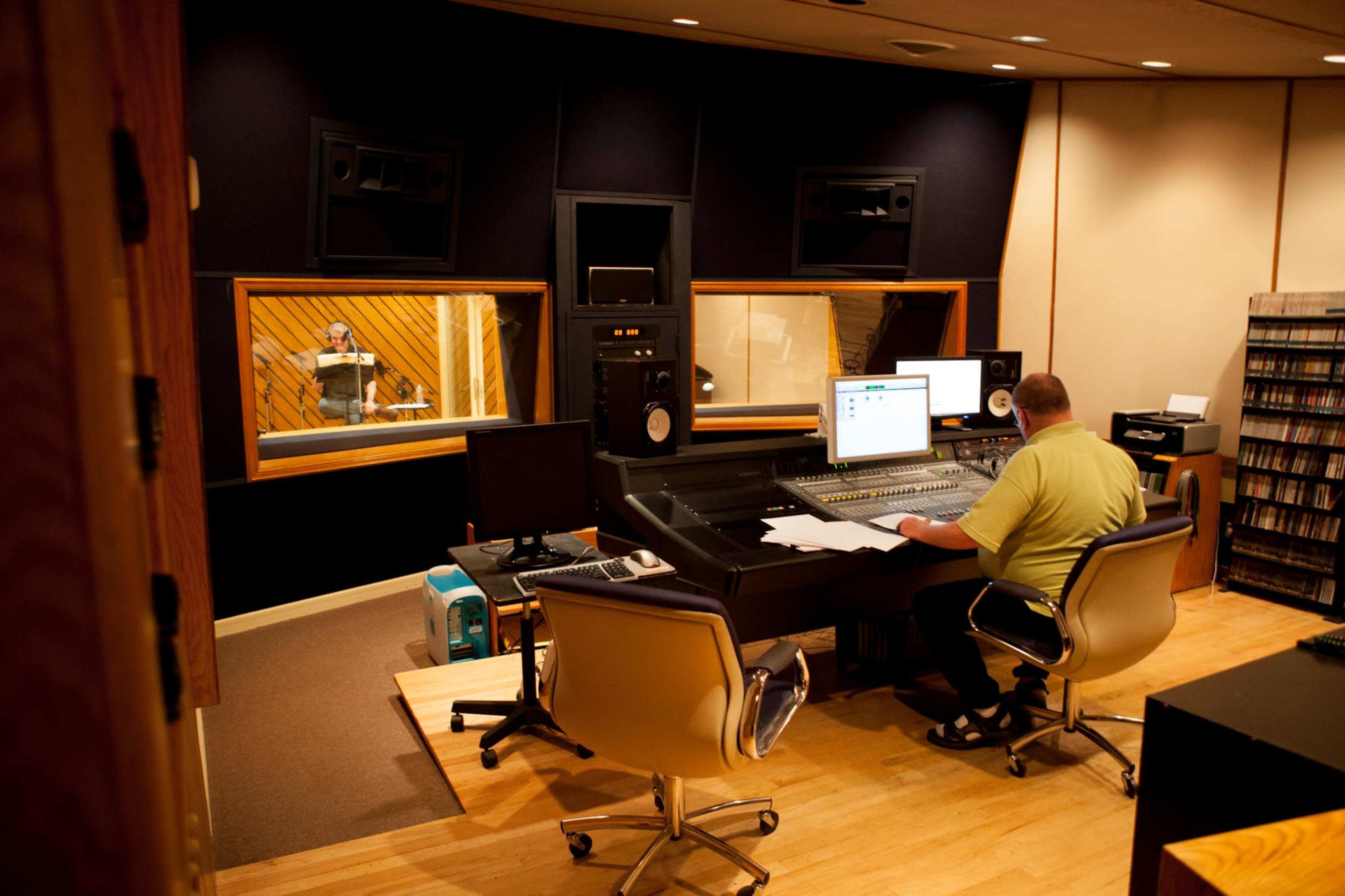 A man sits at a mixing console in a music studio while another person is visible in a recording booth.