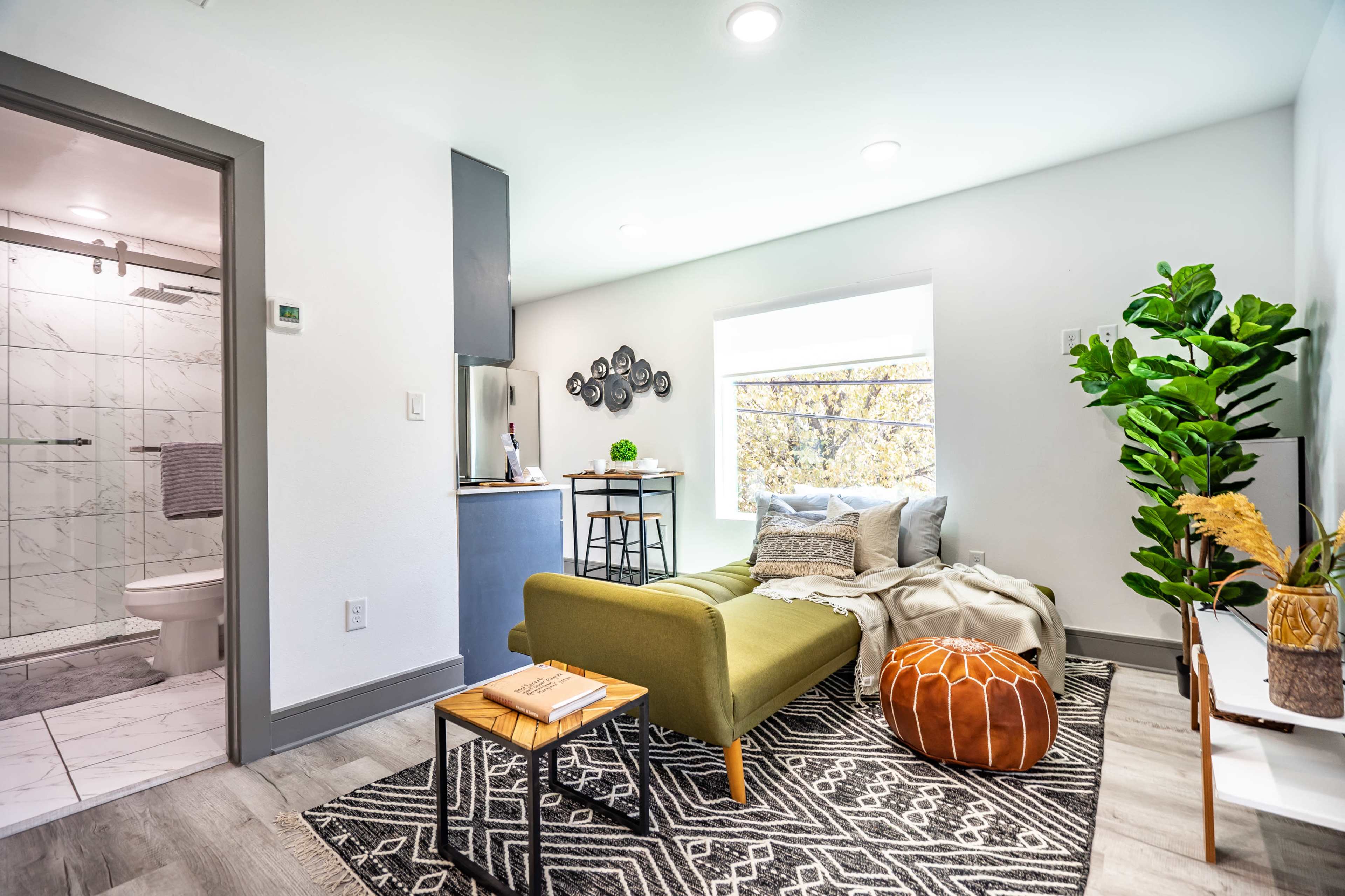 A modern living space featuring a green seating area, a small wooden table, a decorative pouf, and a bathroom visible through an open door.