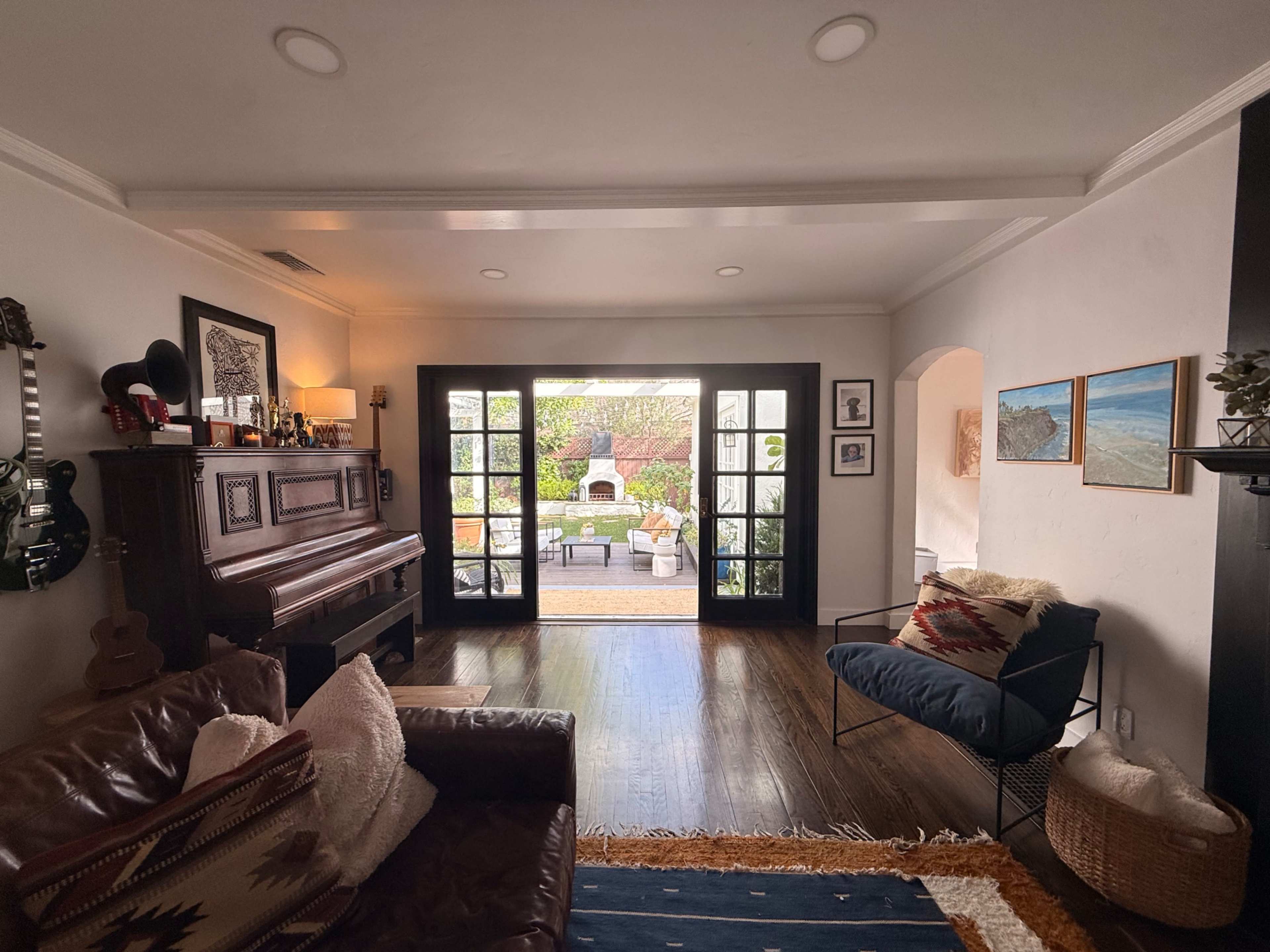 A cozy living room features a piano, a brown leather couch, and sliding glass doors that opening to a landscaped garden area.