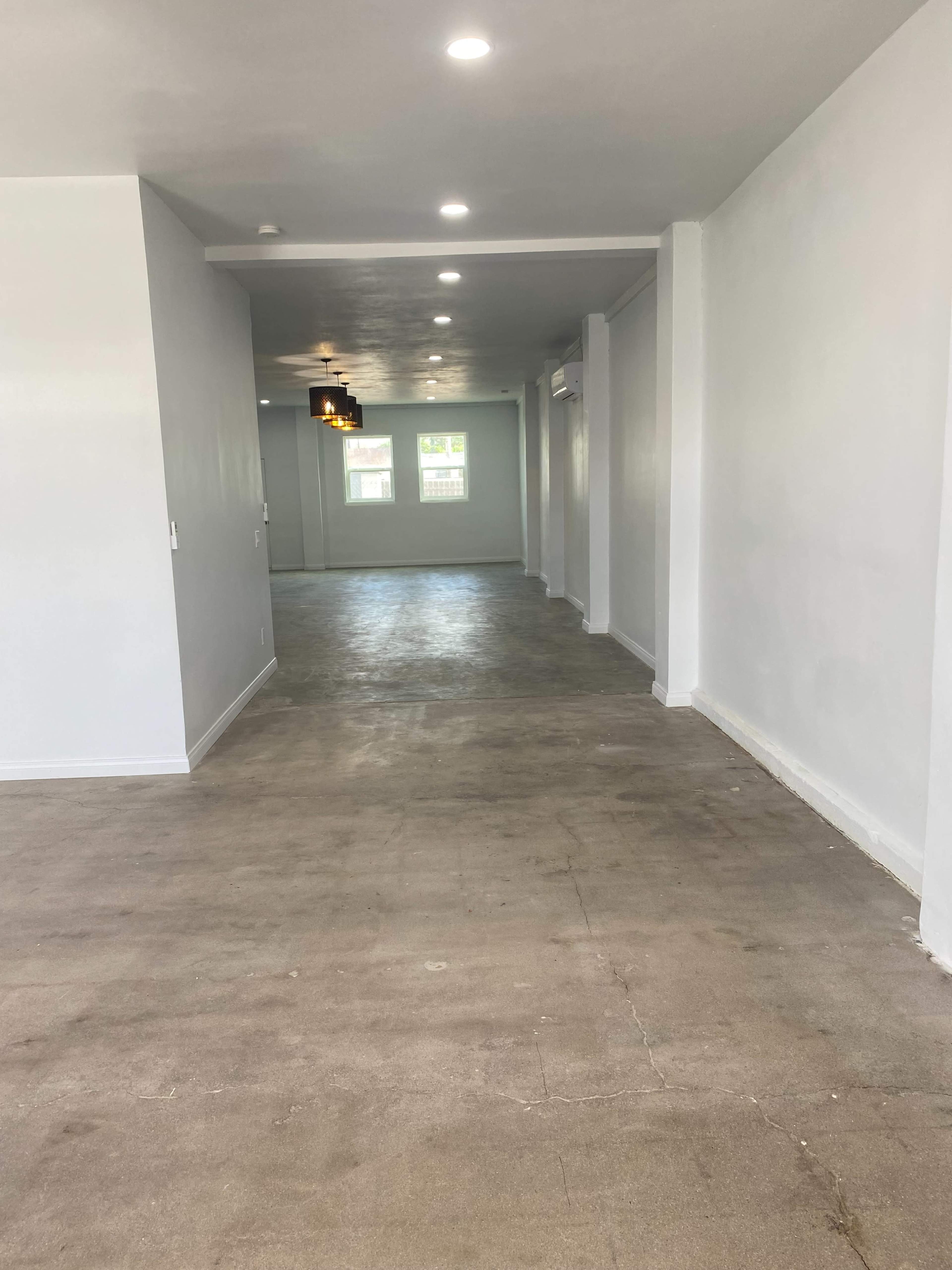 The image shows a spacious, empty interior room with concrete flooring and white walls, featuring recessed lighting and two windows at the far end.