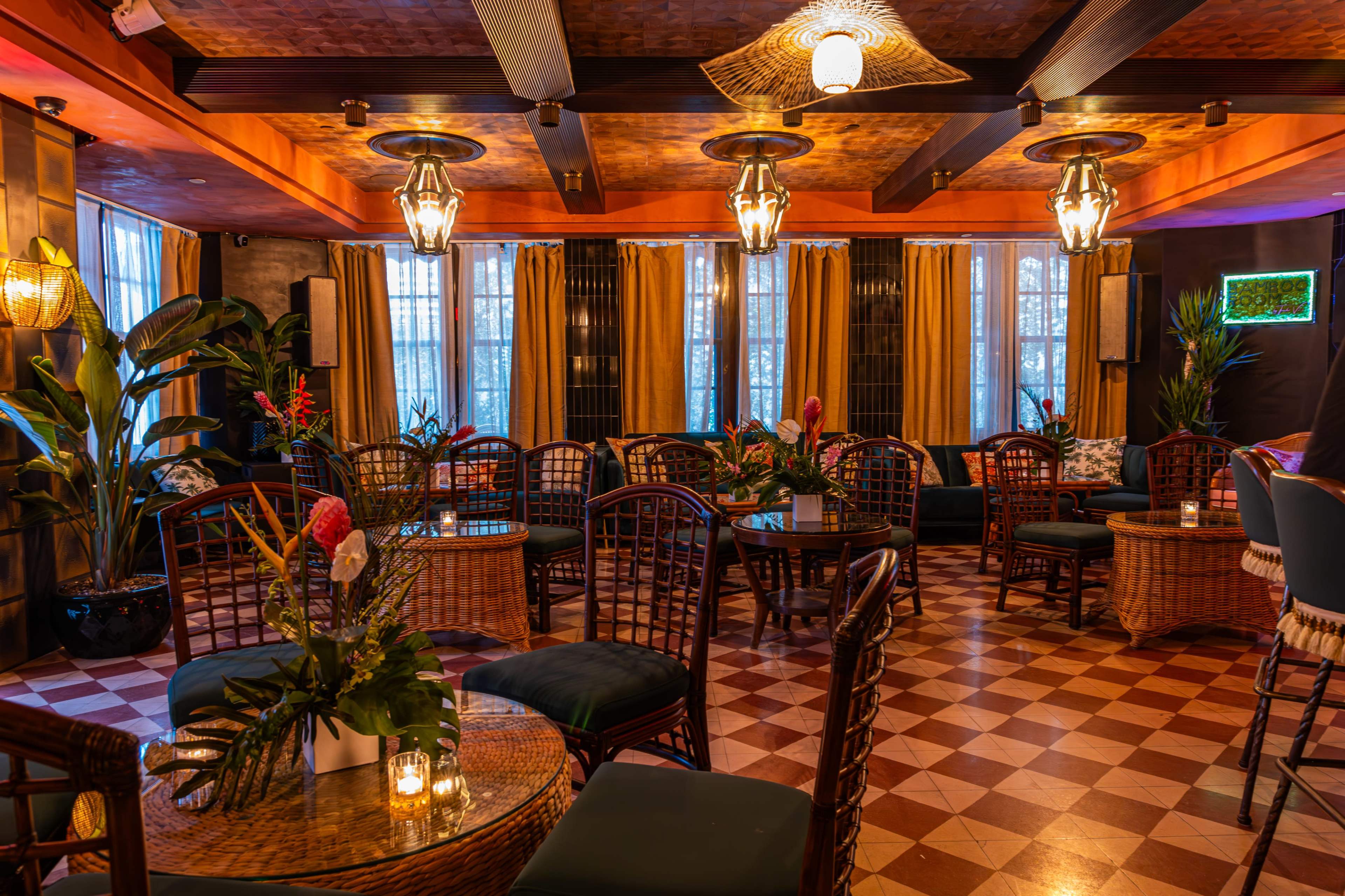 The image shows a warmly lit lounge with wicker furniture, potted plants, and vibrant curtains.