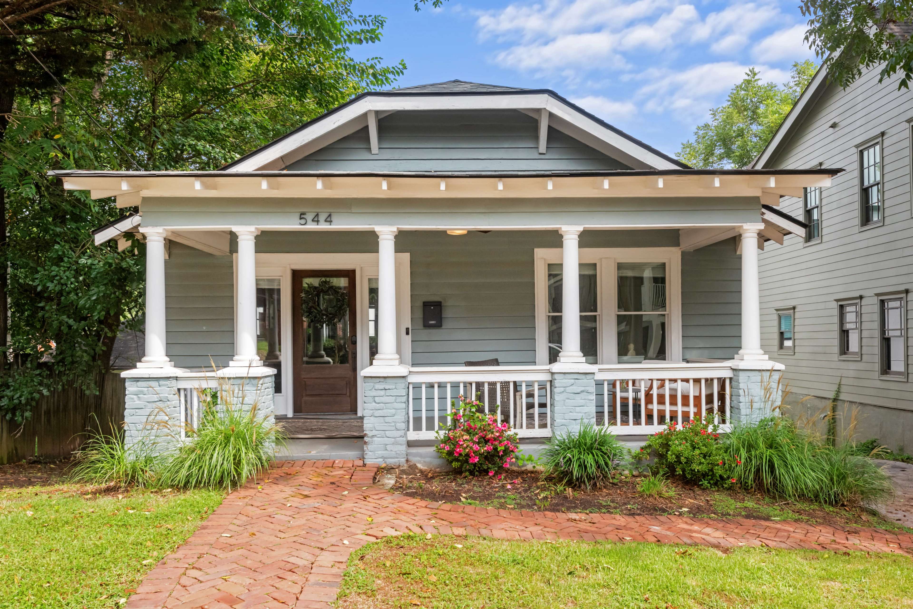 The image shows a light blue, single-story house with a front porch supported by white columns and a brick pathway leading to the entrance.