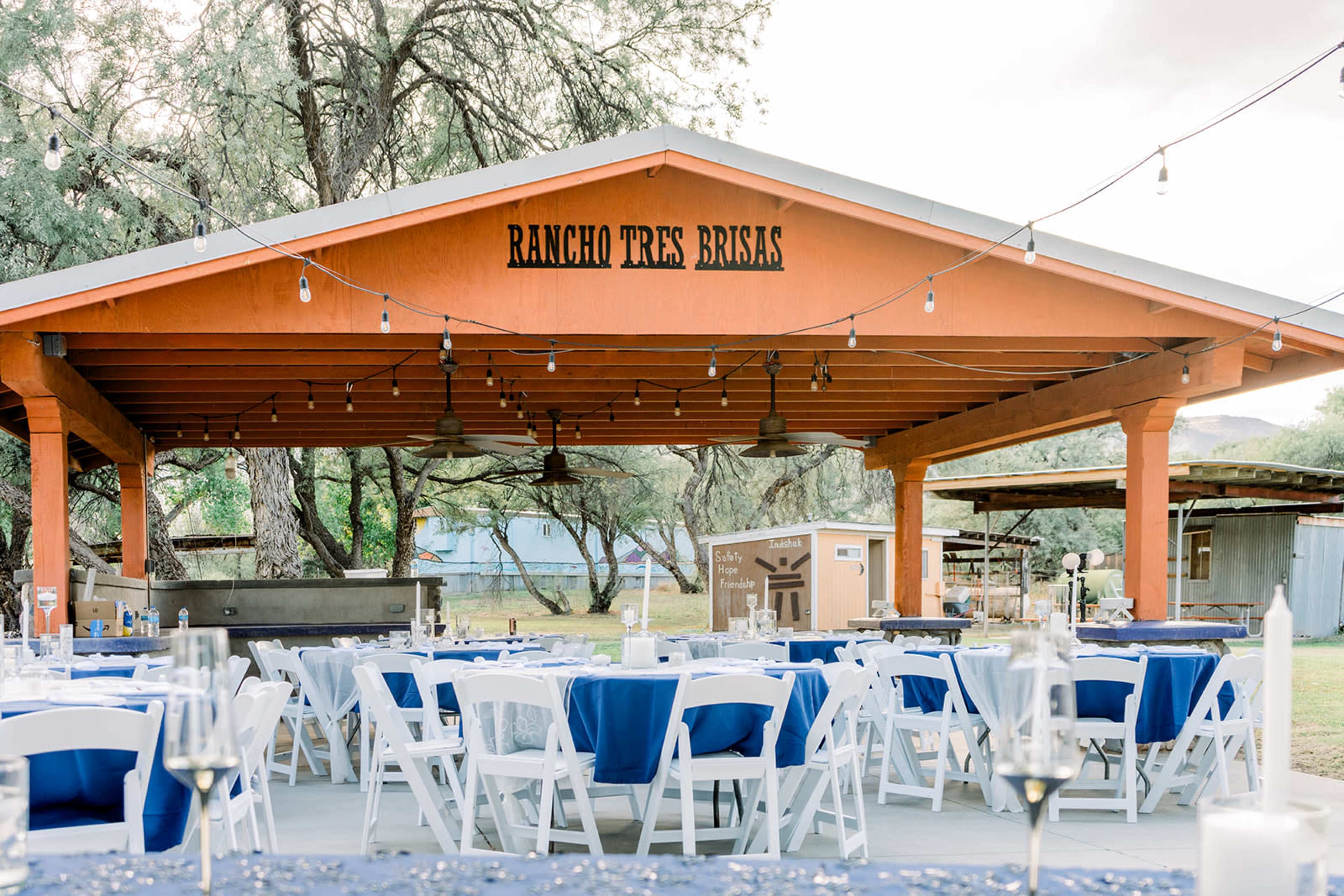 The image shows an outdoor venue with a covered pavilion featuring an orange structure labeled "RANCHO TRES BRISAS," surrounded by tables set with blue tablecloths and white chairs.