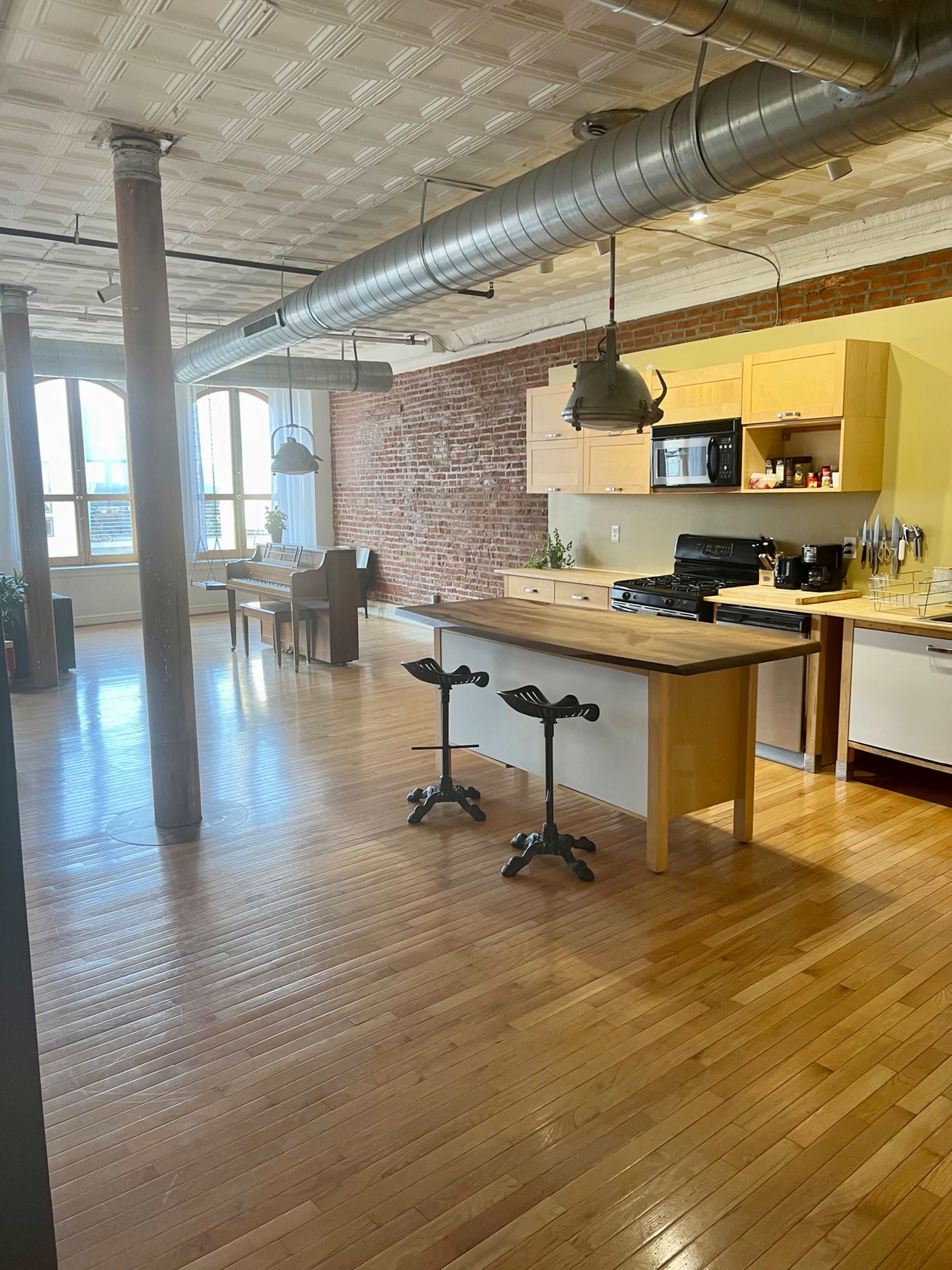 A spacious kitchen with wooden flooring, exposed brick walls, and modern appliances, featuring a central island with seating and large windows.