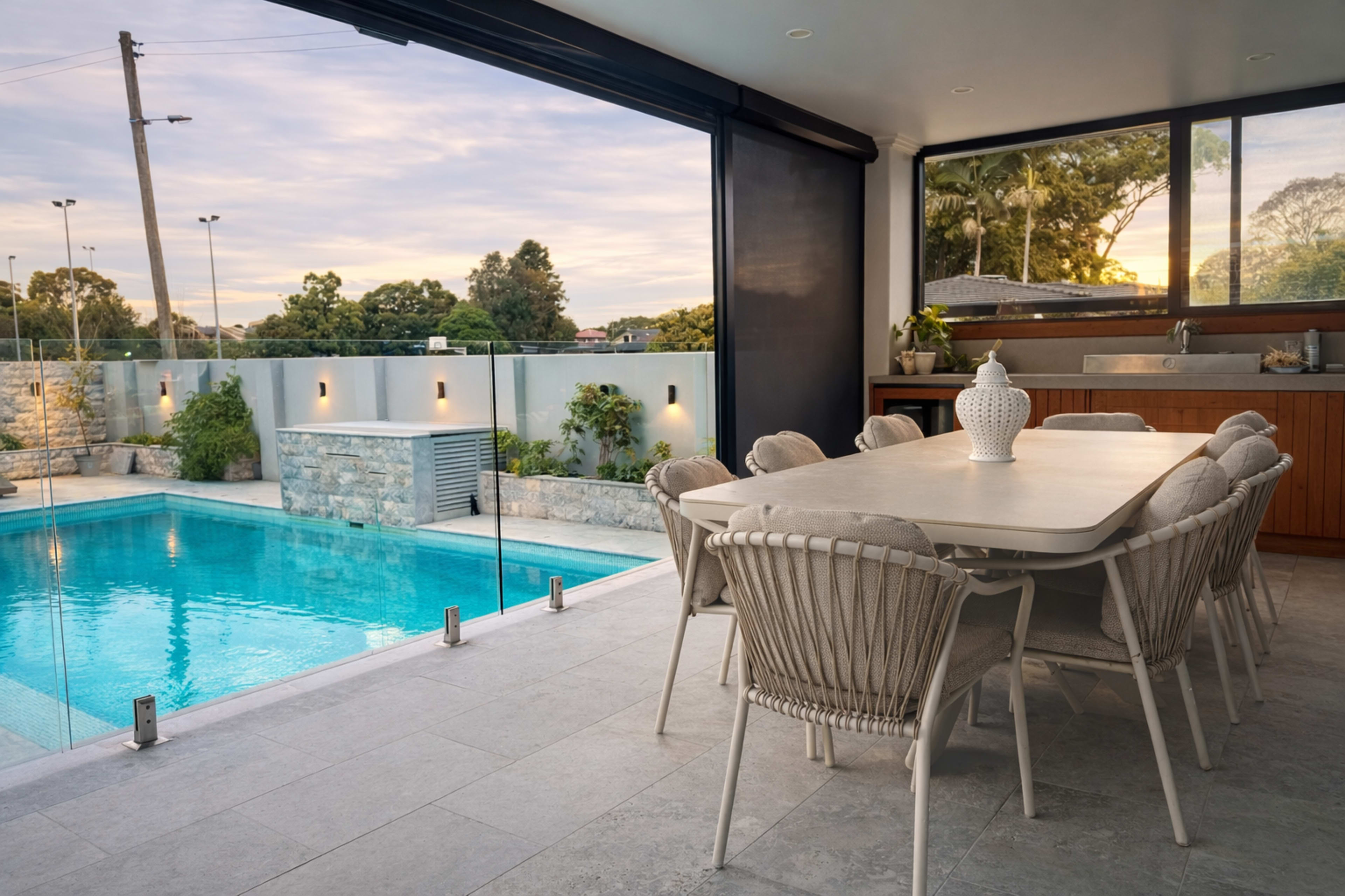 The image shows a modern outdoor dining area adjacent to a swimming pool, with a spacious table surrounded by chairs and large windows revealing a landscaped yard.