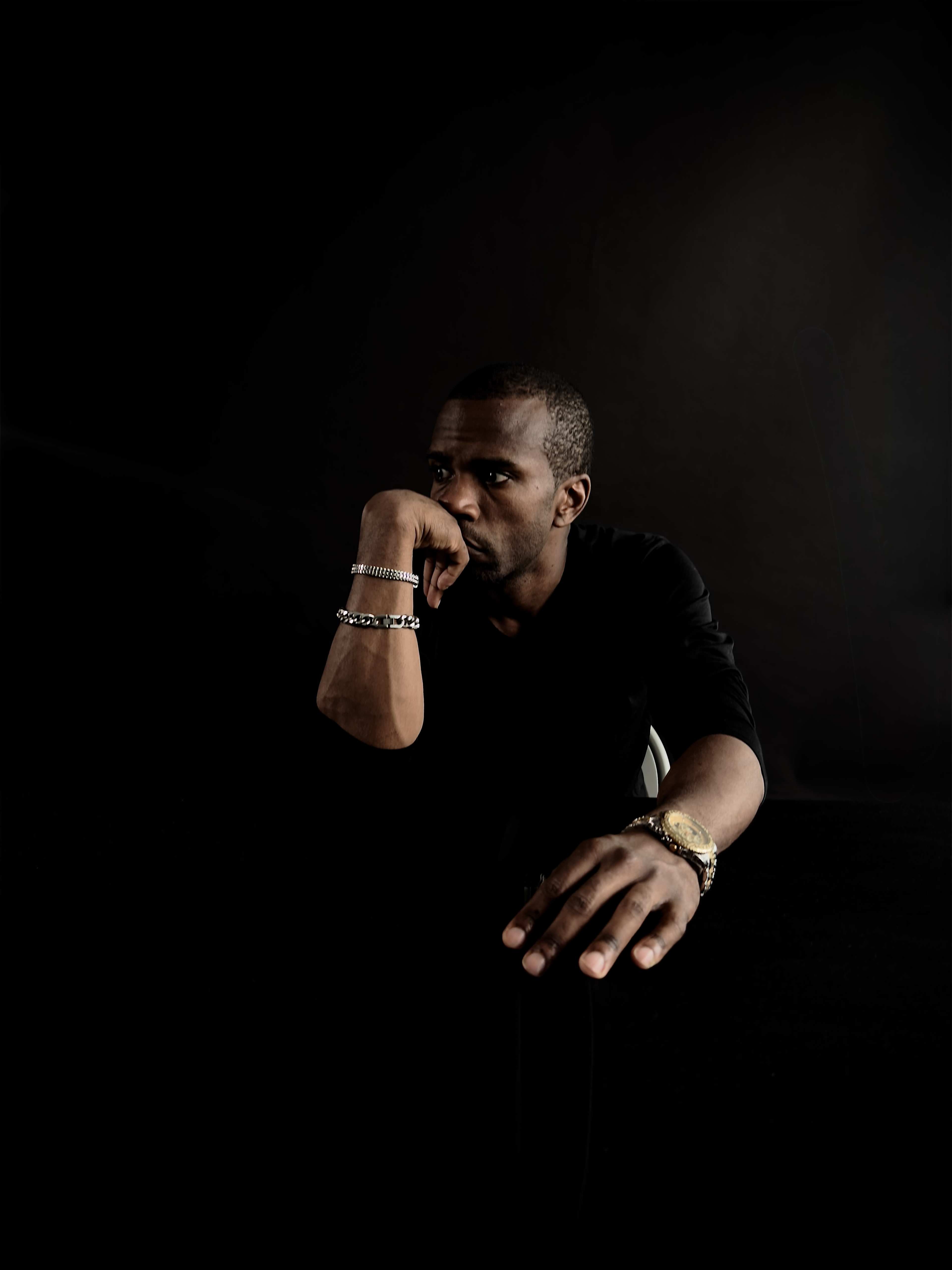 A man rests his chin on his hand while leaning forward, with an outstretched arm against a dark background.