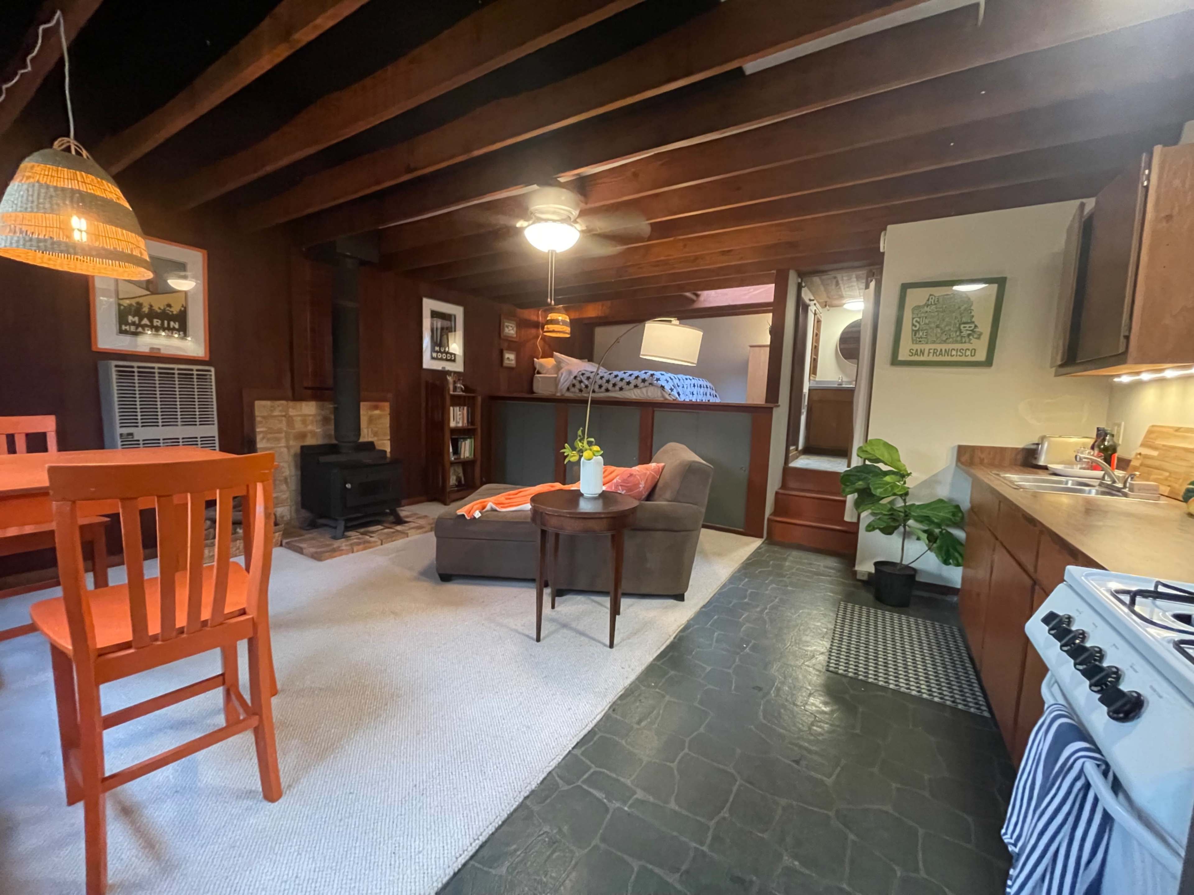 A cozy living area with exposed wooden beams, featuring a seating area with a couch, a dining table, a small kitchen, and stairs leading to a bedroom.