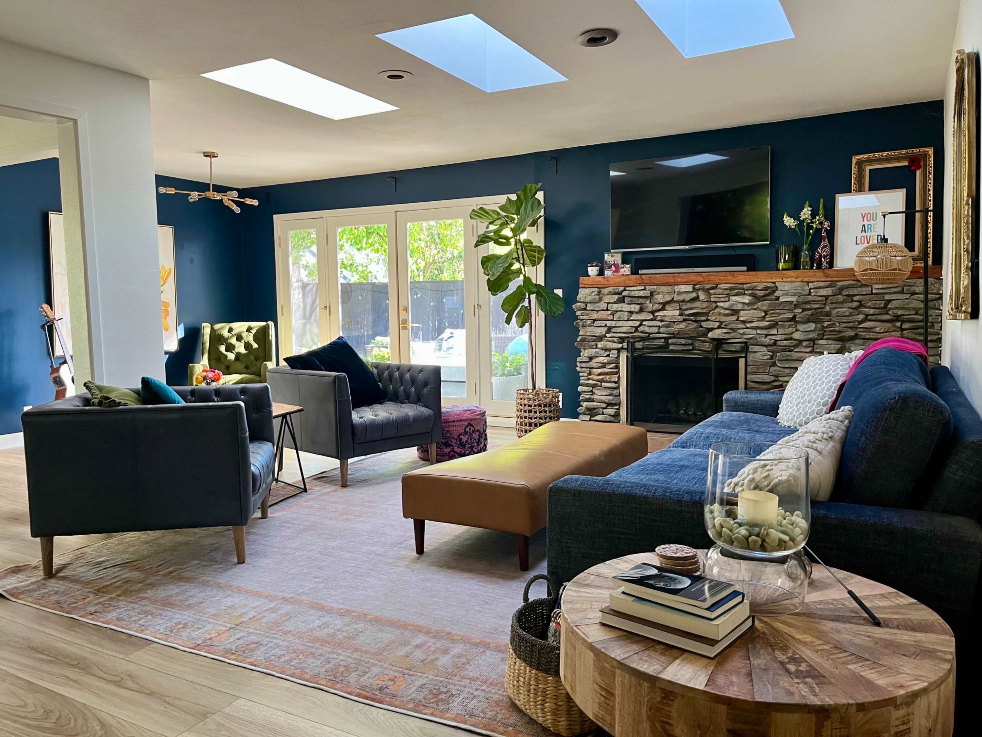 The living room features dark blue walls, large windows with natural light, a stone fireplace, and various seating options including a sofa and armchairs.