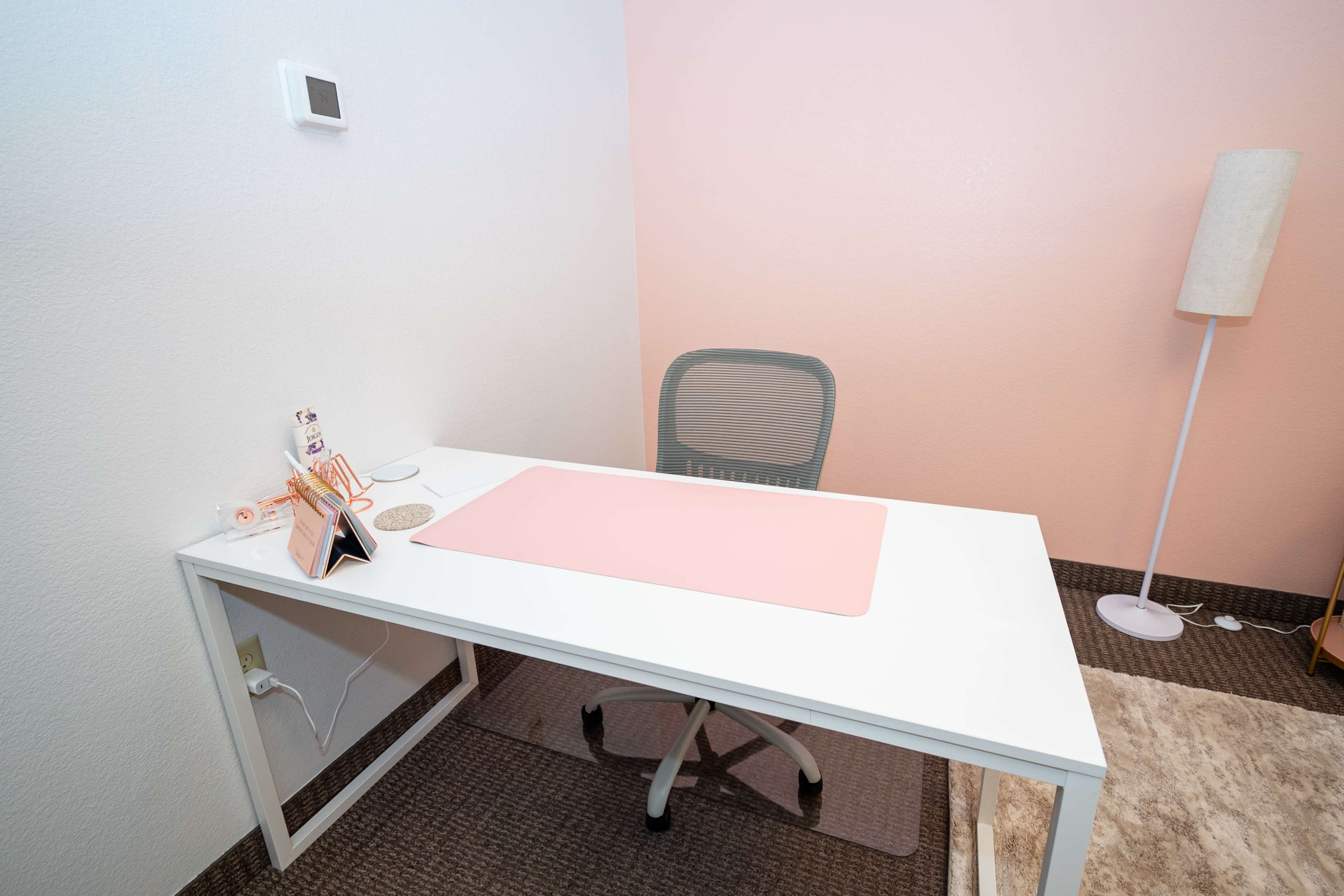 The image shows a minimalistic office space with a white desk, a gray swivel chair, a pink desk mat, and a floor lamp beside the desk in a room with light pink walls.