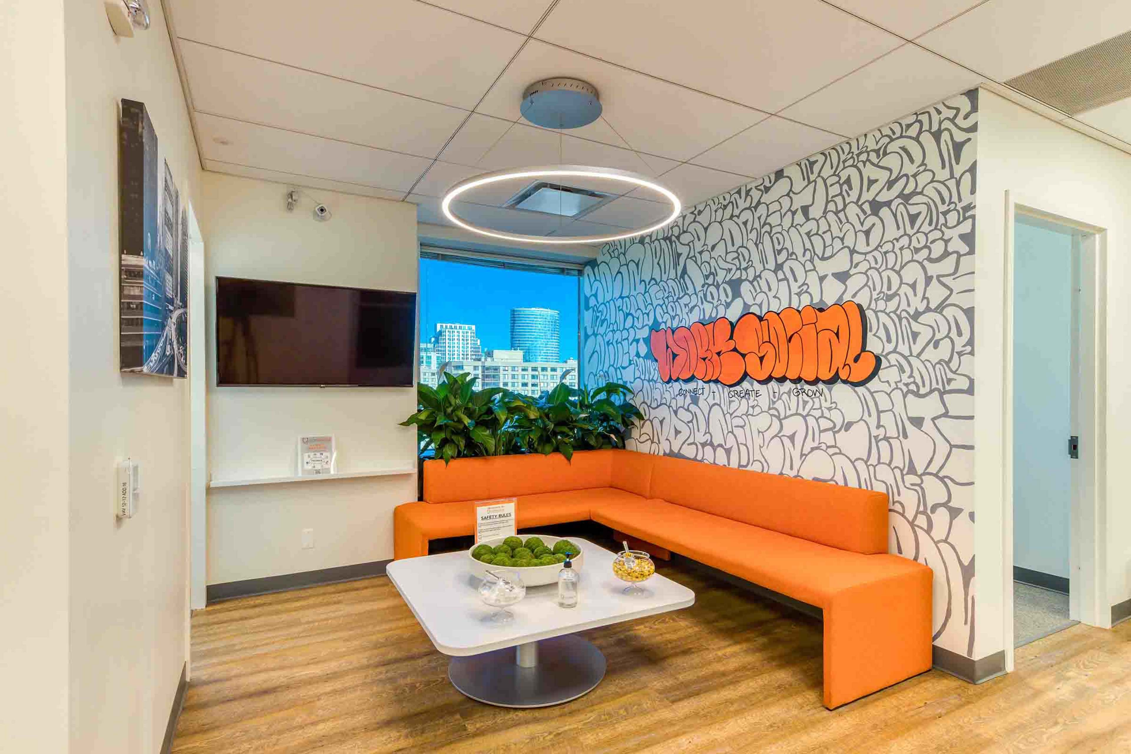 A modern office seating area features an orange sectional sofa, a round coffee table with snacks, and a large window showcasing a city view.
