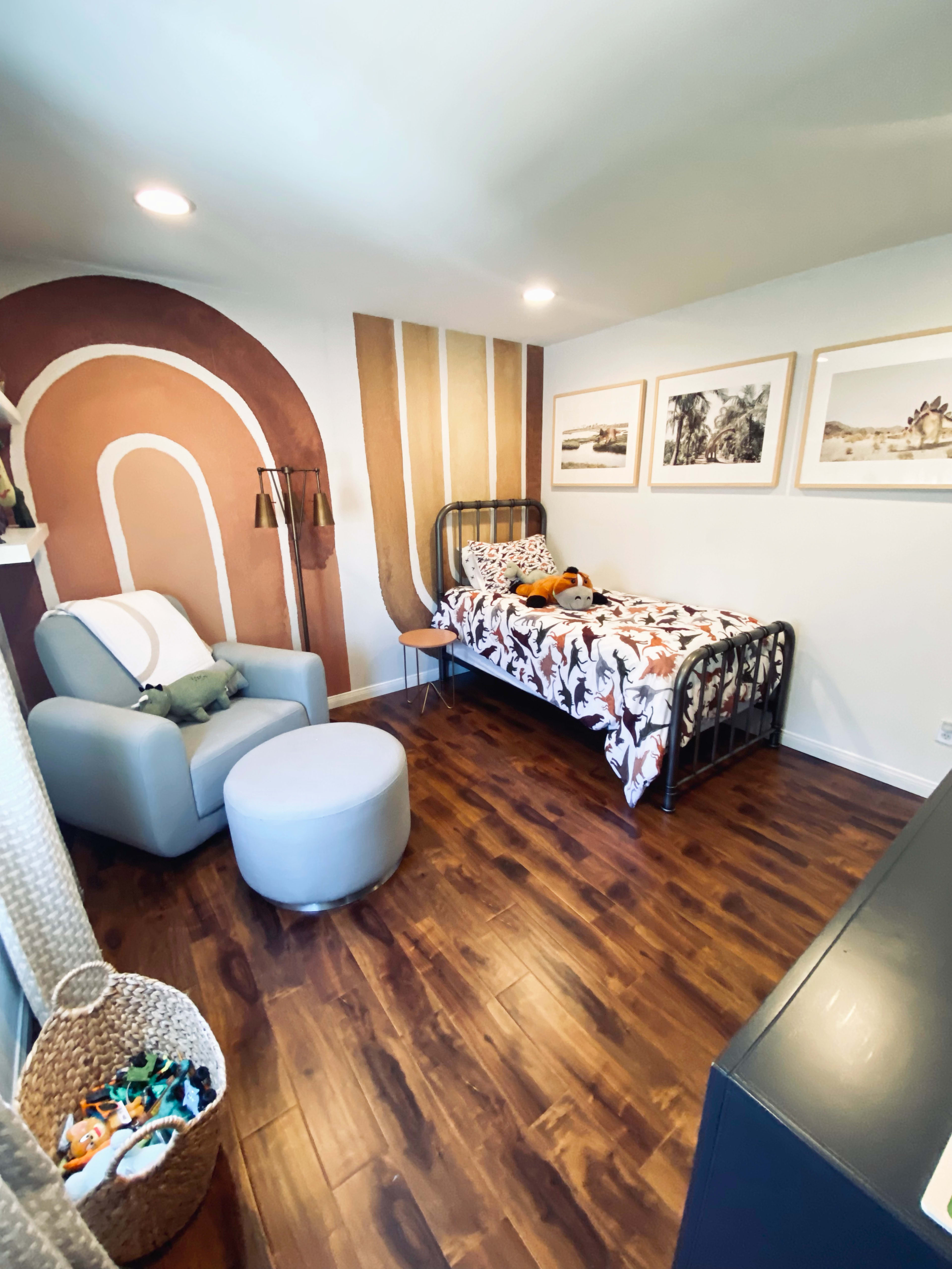 The image shows a children's bedroom featuring a single bed with a floral-patterned blanket, a rocking chair, and artwork on the walls, all arranged on a wooden floor.