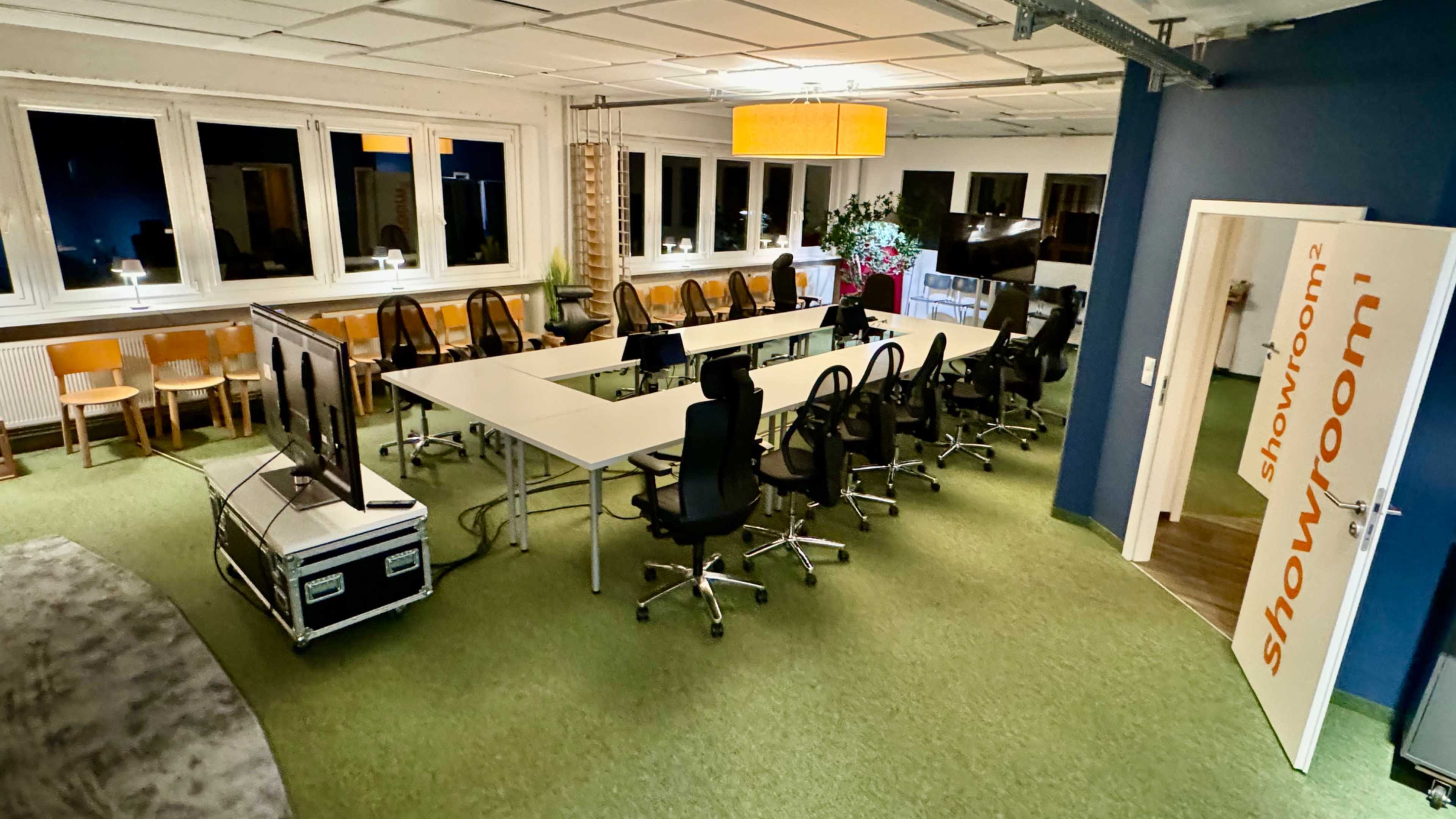 The image shows a spacious showroom with a long table surrounded by office chairs and a large TV at one end, illuminated by overhead lighting.