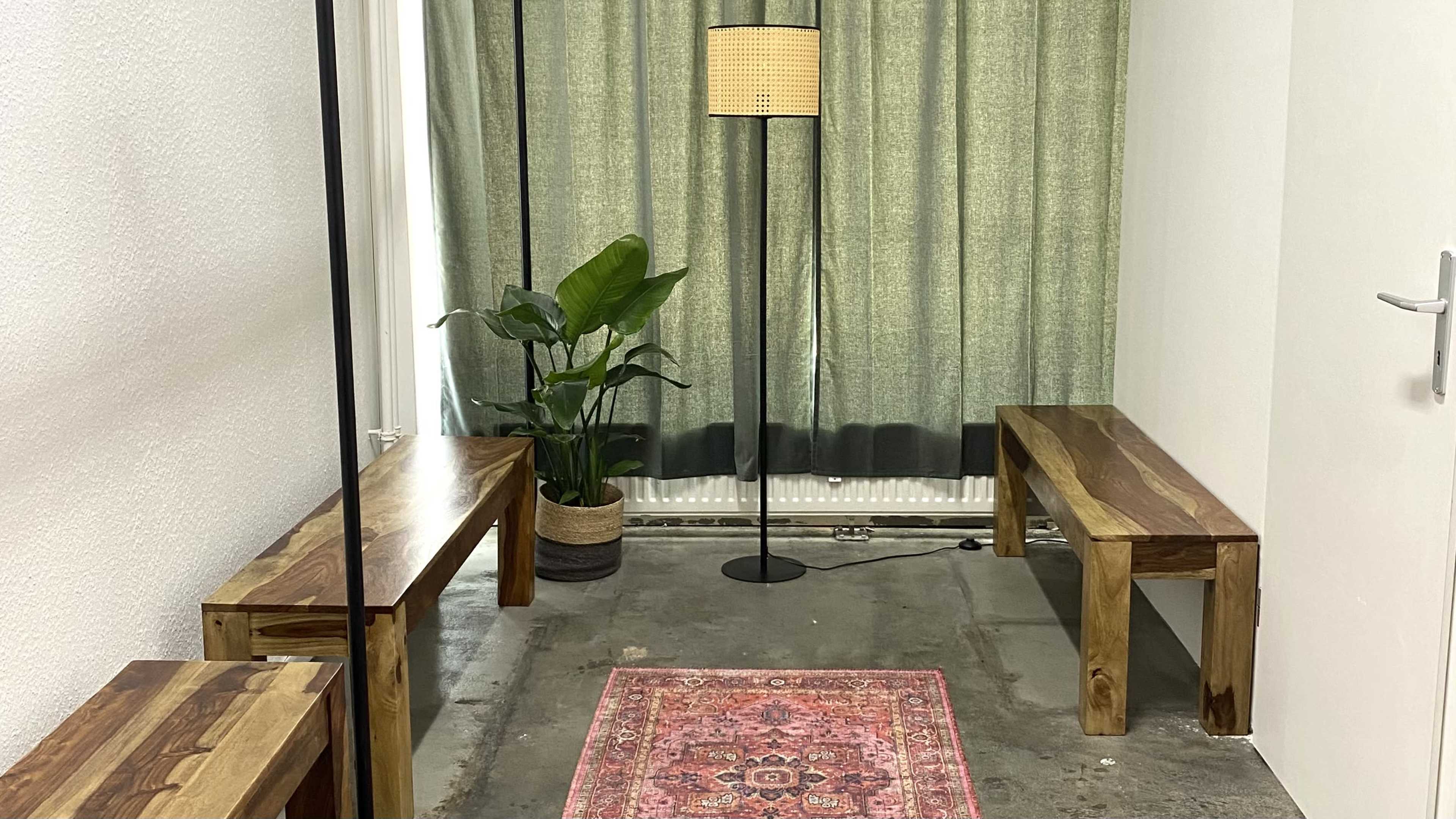 The image shows a small room with two wooden benches, a floor lamp, a potted plant, and a patterned rug on a concrete floor, with green curtains providing privacy at the window.