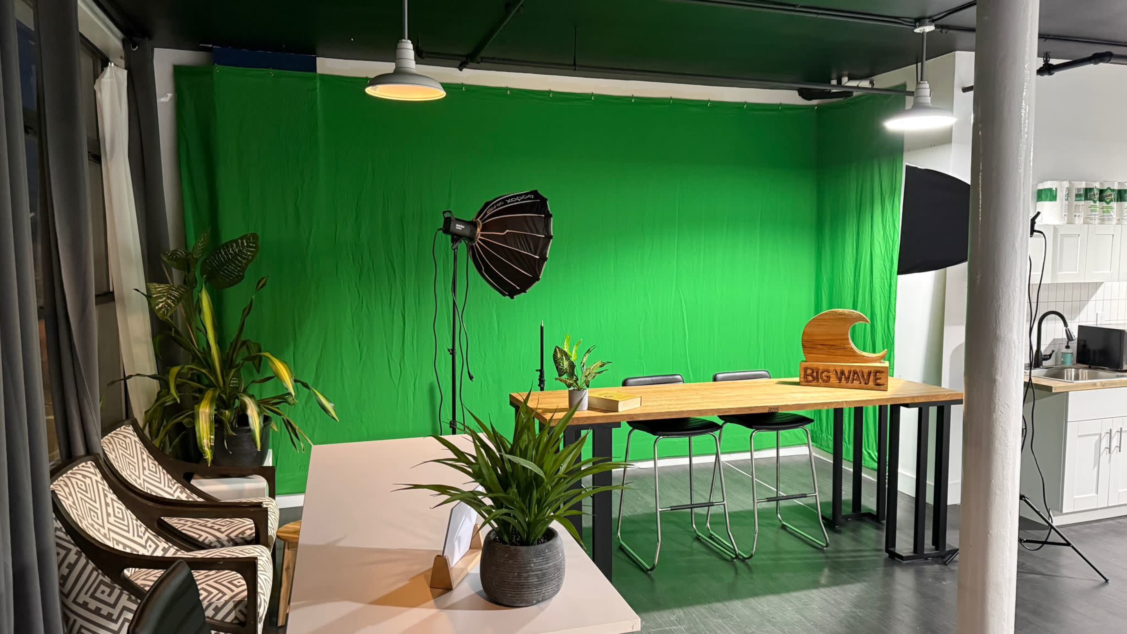 The image shows a well-lit studio space with a green screen backdrop, a wooden table, two black stools, and decorative plants.