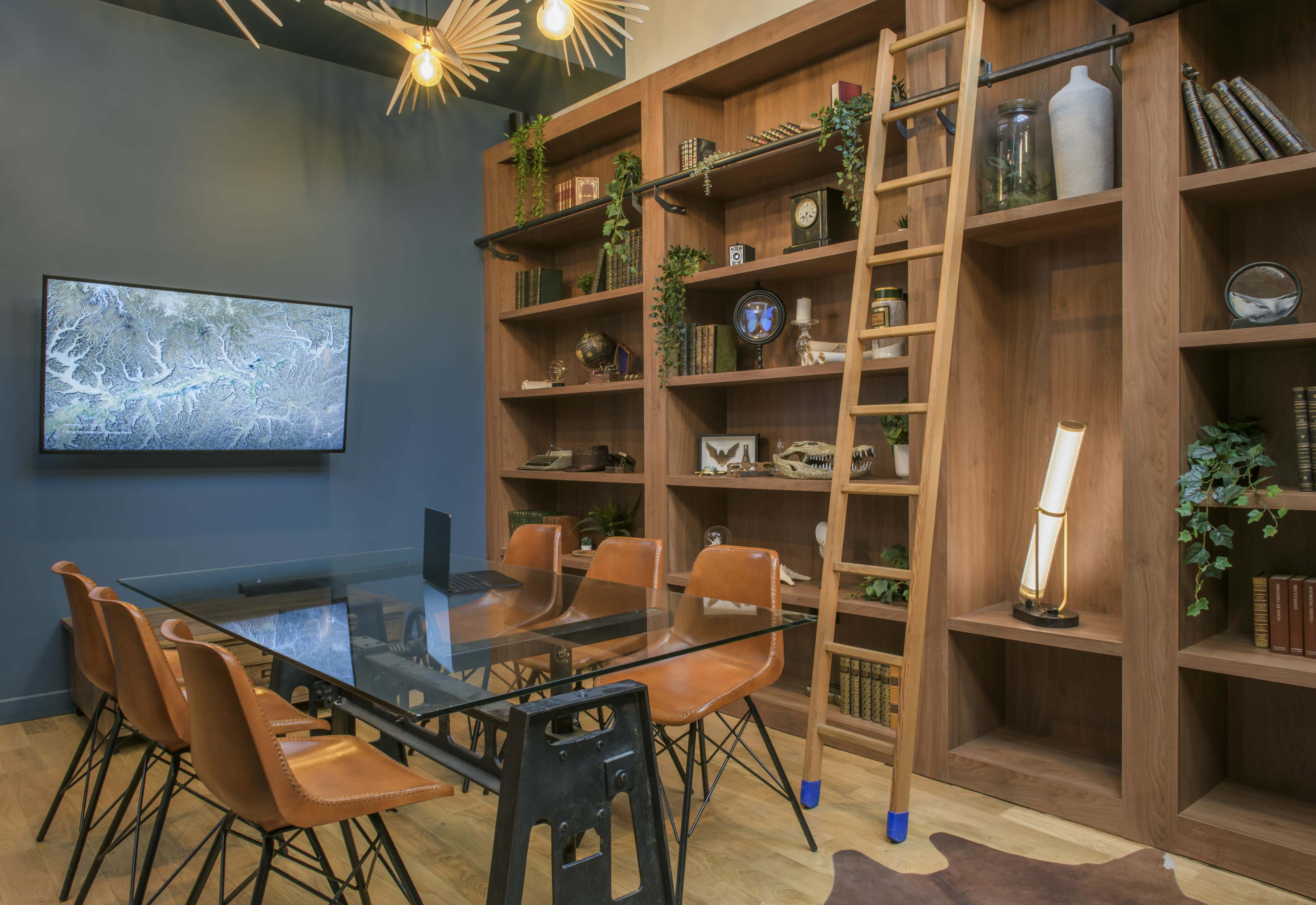 A modern conference room features a glass table surrounded by brown chairs, a wooden ladder leading to shelves filled with various items, and a mounted television displaying a map.