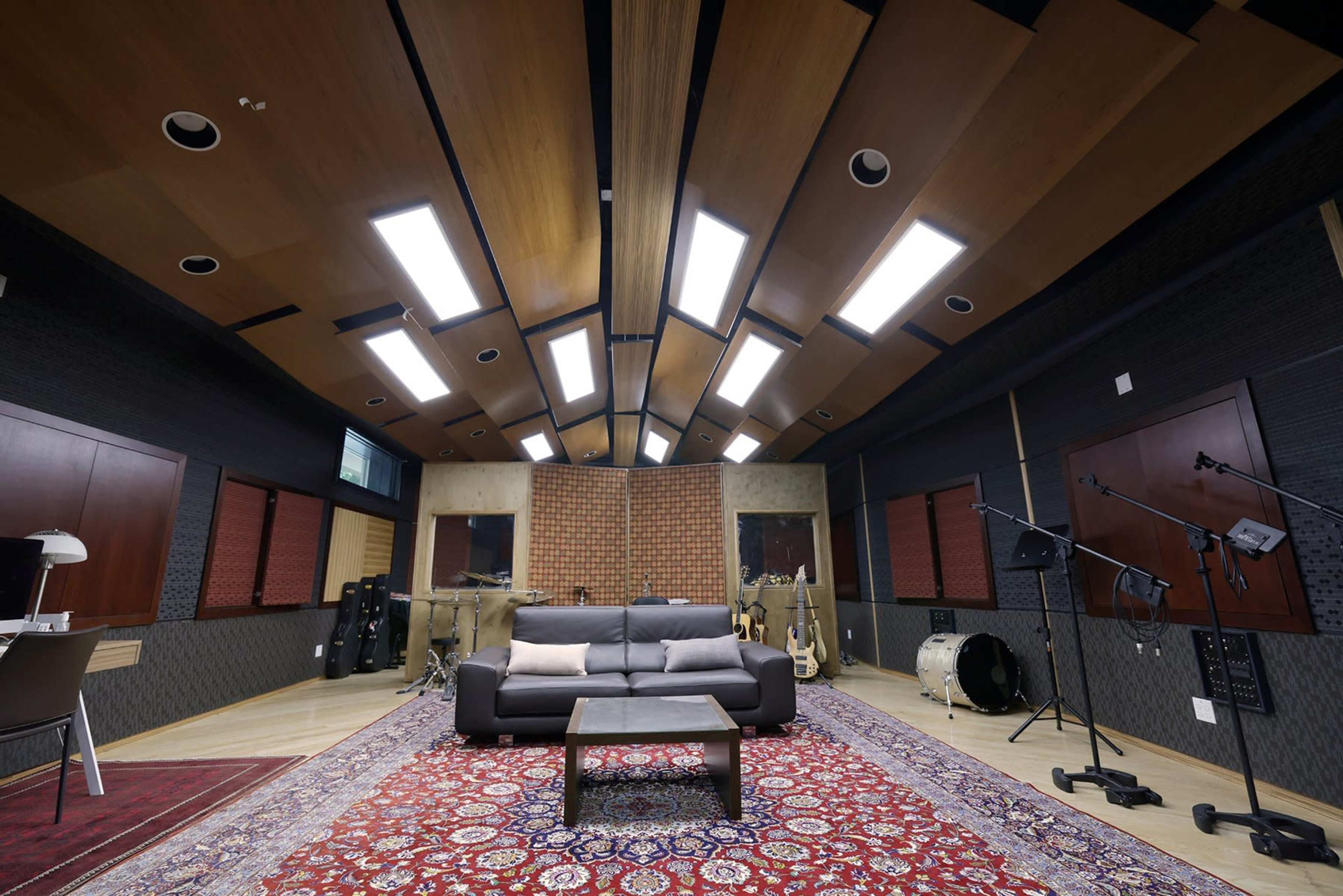The image shows a recording studio with a sectional sofa, a coffee table, and various musical instruments arranged around a patterned rug.