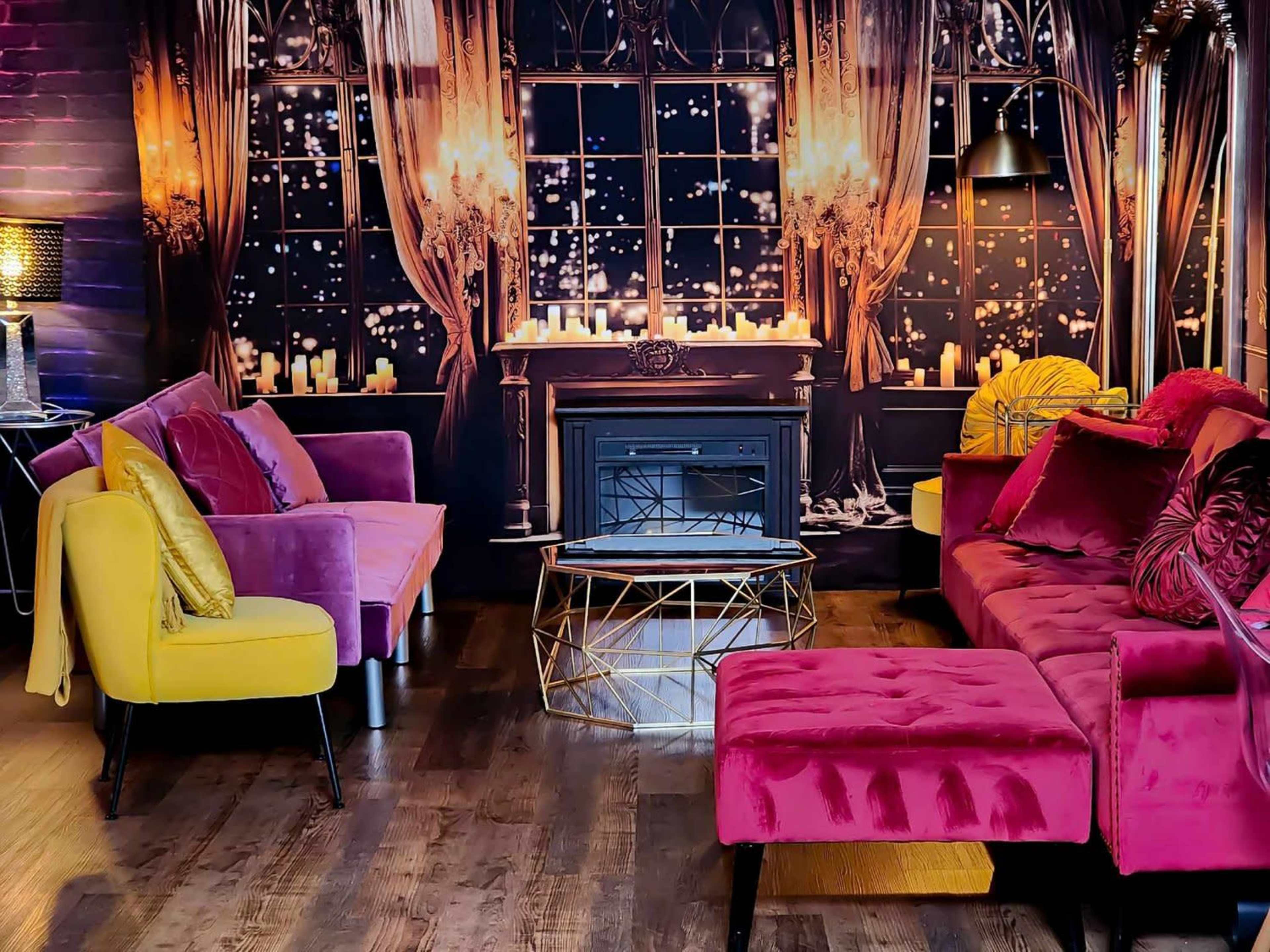 A stylish living room with pink and yellow couches, a black fireplace, and large windows decorated with curtains and candles.