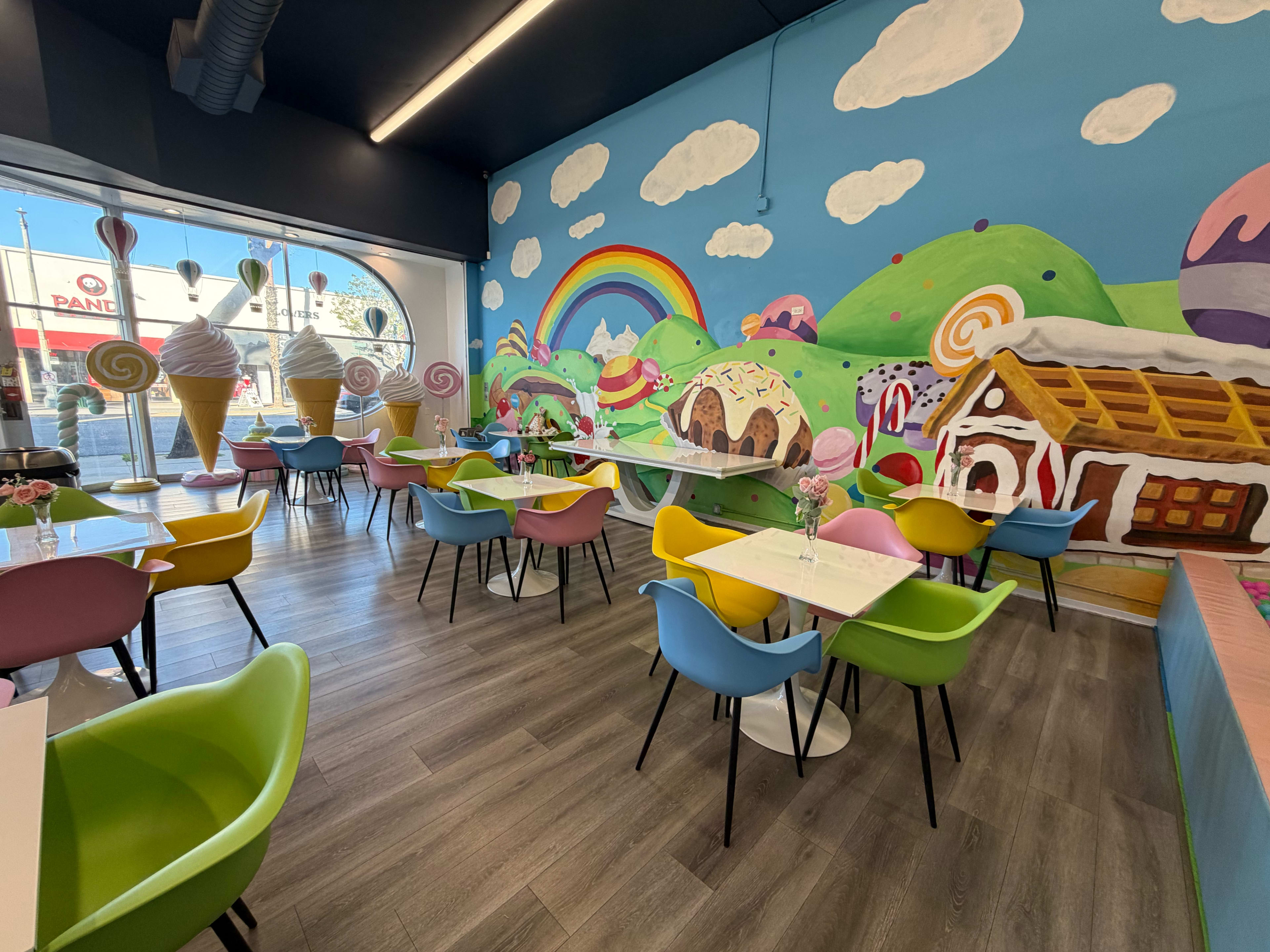 The image shows a vibrant ice cream shop interior with colorful seating, tables, and a whimsical mural featuring ice cream cones, clouds, and a rainbow.