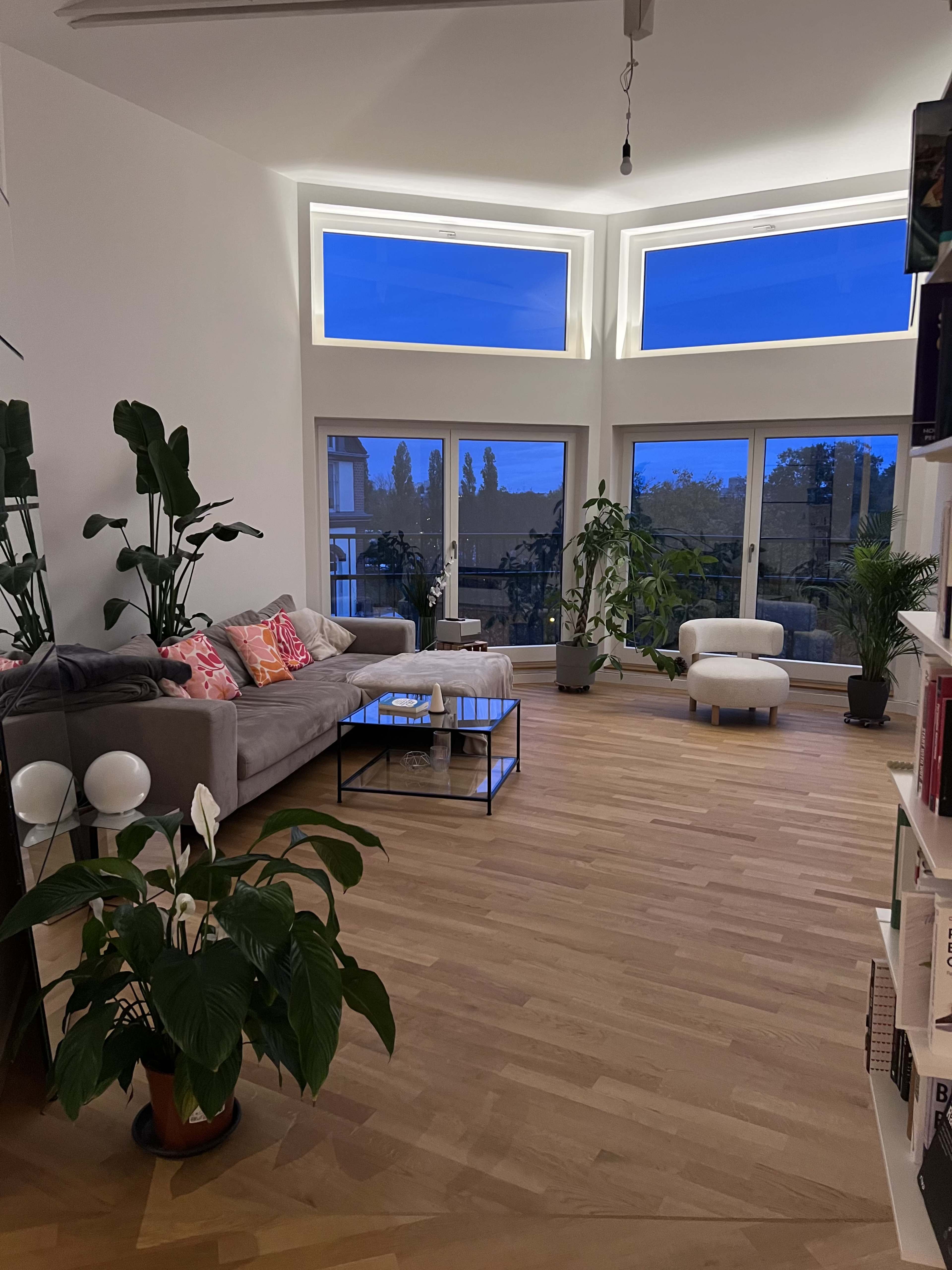The living room features a sectional sofa, a coffee table, and large windows that overlook a garden area, all complemented by various indoor plants.