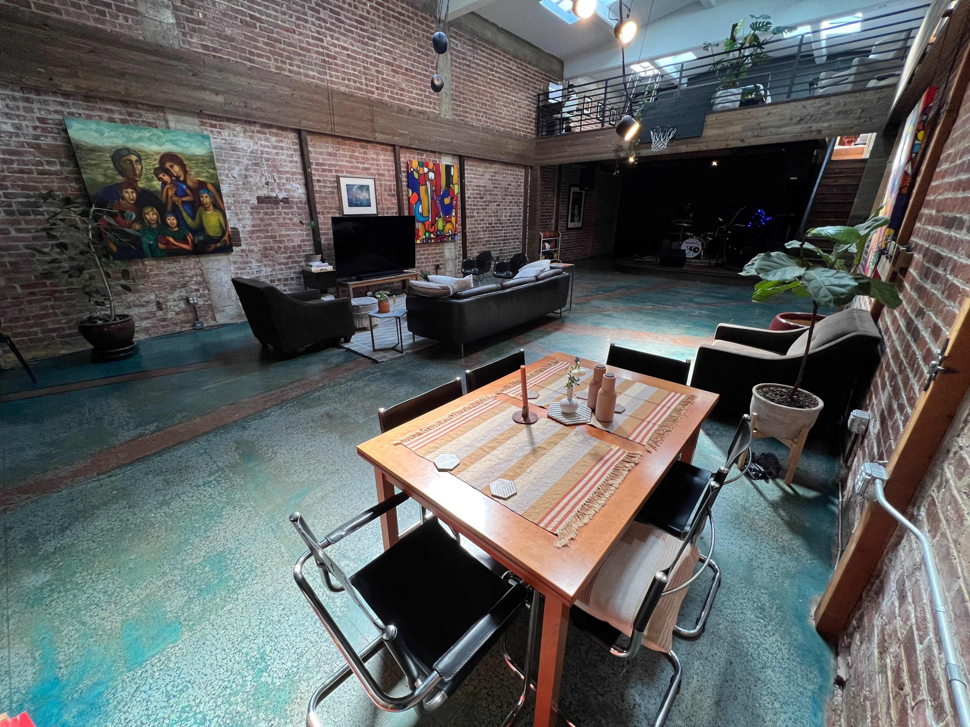 An open loft space featuring exposed brick walls, a dining table with candles, a sofa area, and a stage with musical instruments in the background.