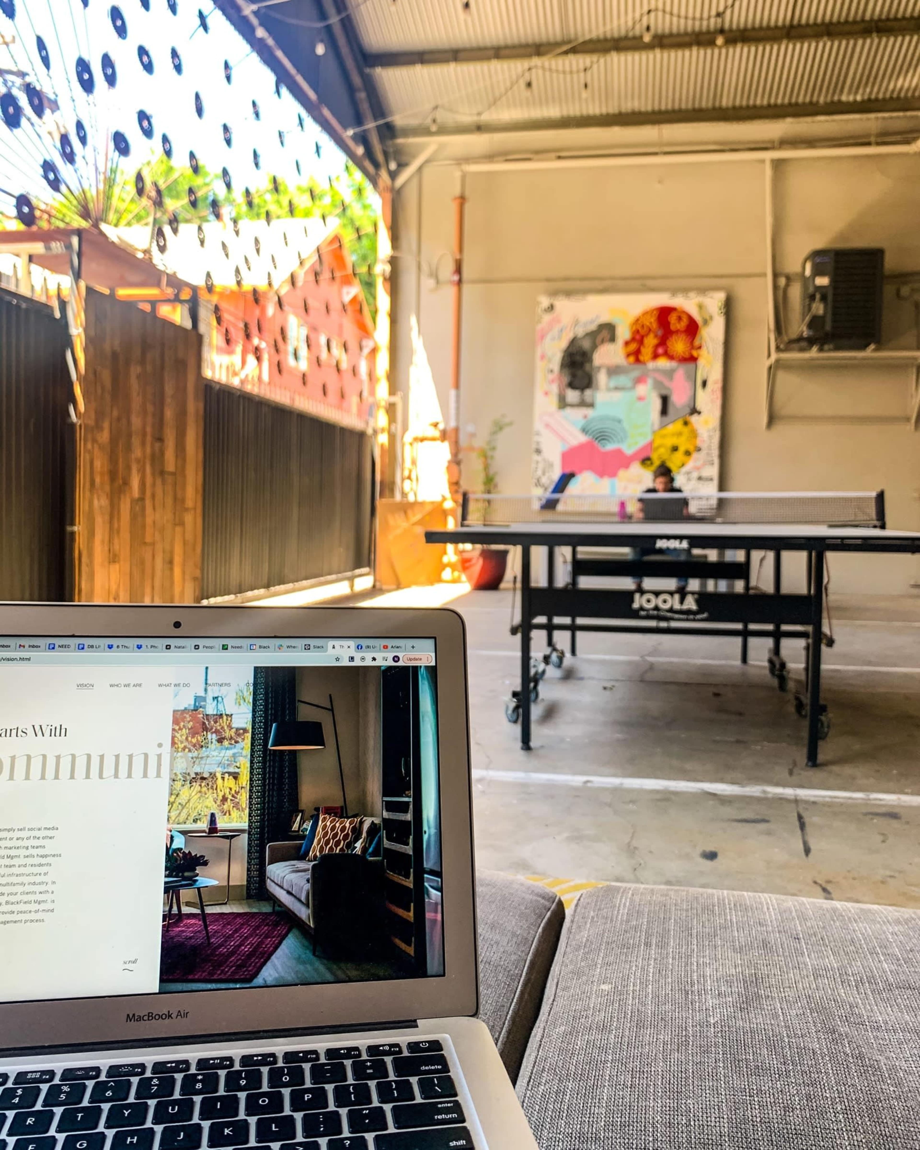 A laptop on a couch in a spacious, well-lit room with a ping pong table and colorful artwork on the wall.